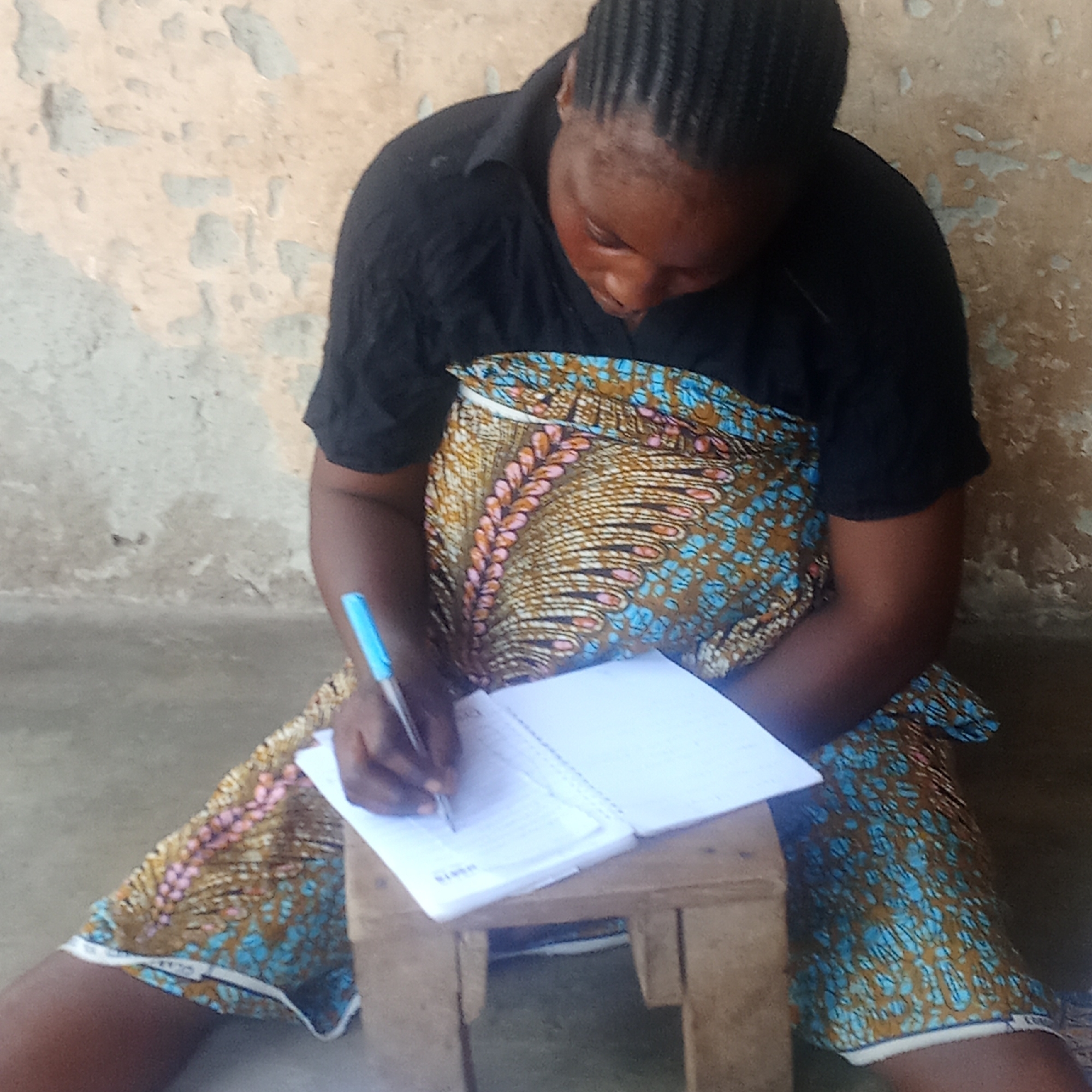 A medium shot captures an individual, likely a woman, sitting on the floor and writing in a notebook in an indoor setting in Akwanga, Nigeria. She is clad in a dark, plain short-sleeved top and a colorful patterned wrapper featuring designs in brown, blue, pink, and white. Her dark hair is styled in braids.
The individual is leaning forward, intently focused on writing. She holds a blue ballpoint pen in her right hand, actively putting it to paper in an open, spiral-bound notebook. This notebook is supported by another notebook underneath it, both resting on a small, rectangular wooden stool that acts as a makeshift desk. On one of the notebook covers, the word "SCHOOL" is partially visible on a white label. Inside the notebook, some written text is discernable, possibly including the word "NIMES" or "SIMES".
The background features a light-colored wall that exhibits a worn, textured appearance with areas of peeling paint or plaster. The floor is a simple, light-colored surface. The natural lighting suggests it is daytime. The overall scene depicts focused activity, possibly studying or note-taking, in a modest environment.
