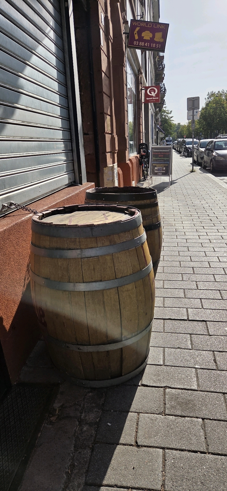 The image depicts a street scene in Strasbourg, France, on a sunny day. In the foreground, two wooden barrels are positioned on a brick-paved sidewalk. The barrel on the left is partially in shadow, while the one on the right is more fully illuminated by sunlight. Behind the barrels, a building with red brick facade is visible, featuring a silver roll-up door on the left and shop windows. A sign with "WORLD LINK" is affixed to the building above the windows. Further down the street, a row of parked cars lines the right side, and a bicycle with a basket is leaning against the building. An A-frame sign on the sidewalk advertises services like "RESTAURANT TELEPHONIE," "Impression Photocopie," "scan," and "Transfert d'argent." The sky is clear and blue.