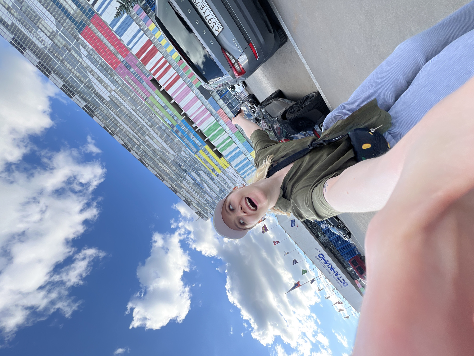 A rotated, wide-angle selfie shows a fair-skinned person with blond hair, wearing a green shirt and grey baseball cap, pointing toward a gray Škoda Kodiaq car parked on a concrete surface. Behind the car, there is a modern building with colored stripes (red, pink, blue, green, yellow) on its facade. Part of the person's lower body, wearing blue striped pants, is visible in the lower right. The sky above features a mix of blue and white clouds. To the right, there is a structure with flags and the text "Международный технический центр АО «НПО Энергомаш» имени академика В.П. Глушко" which identifies the location as Khimki, Russia. A black bag is on the ground beside the person.