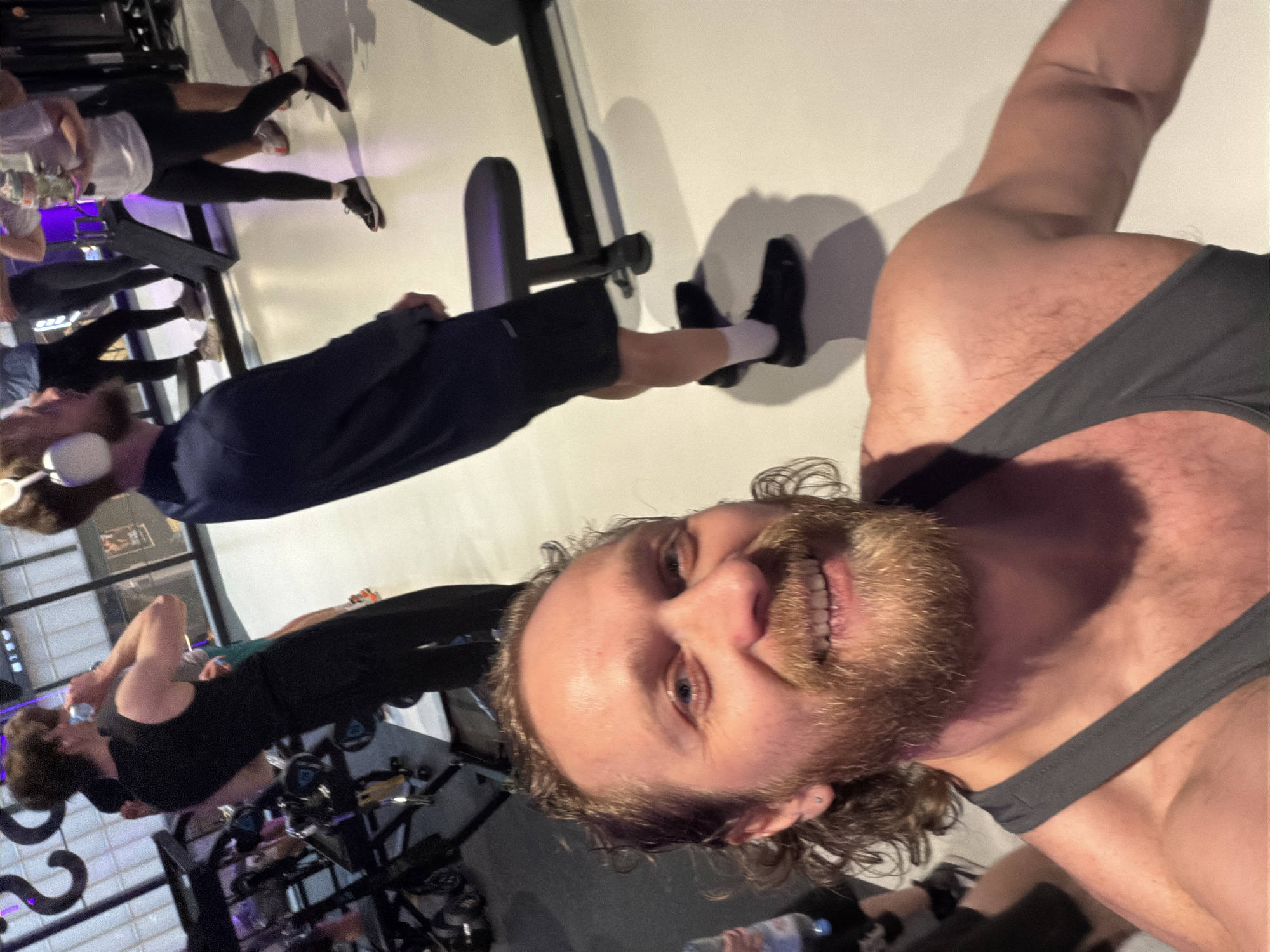 The image is a selfie taken by a man with a smiling expression, a full beard, and curly brown hair, wearing a gray tank top. He has blue eyes and an earring in his left ear. His upper arm and shoulder are visible, showing some body hair.
The setting appears to be a fitness studio or gym in Saint Petersburg, Russia, where a group fitness class, likely indoor cycling or spinning, is taking place. In the background, several other individuals are visible, engaged in the activity. They are positioned on stationary bikes, wearing athletic wear including leggings, shorts, and tank tops, along with sneakers. One person closer to the foreground is wearing a dark blue hoodie and white headphones, standing near a bike. Another participant is seen in a black tank top further back.
The room has white walls and bright artificial lighting, casting shadows from the people and equipment on the wall. The atmosphere suggests an active and dynamic workout session. A water bottle with the brand name, possibly "Evian," is partially visible on the floor in the bottom right corner of the image.
