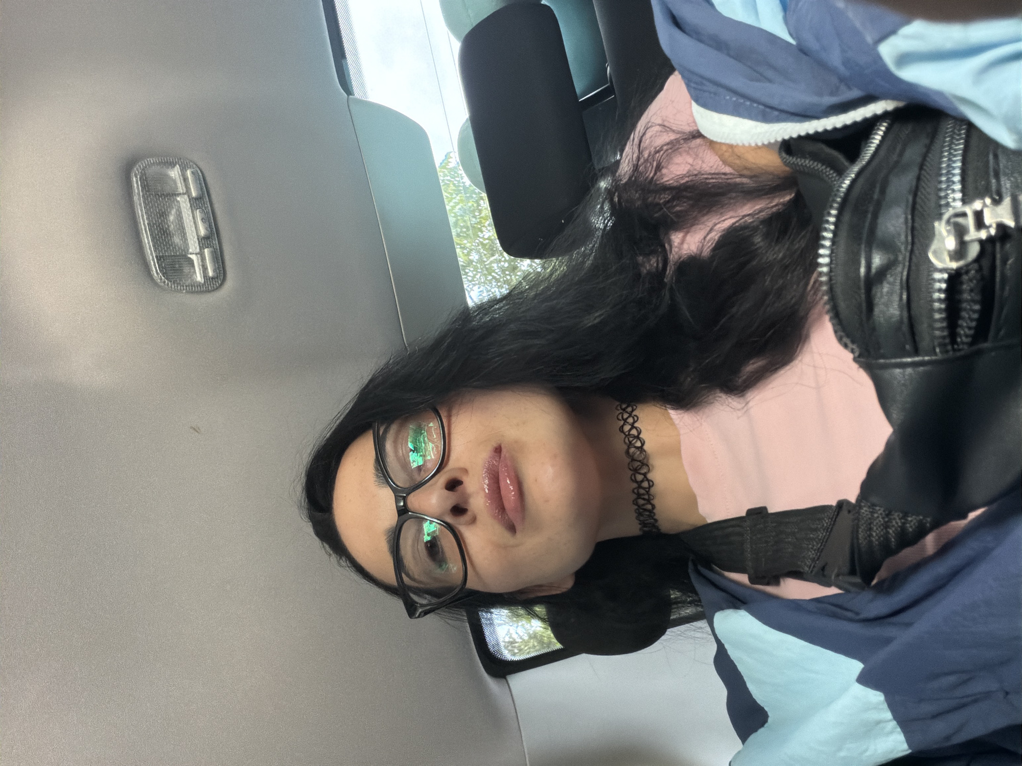 A young woman with dark hair and glasses is in the backseat of a car. She is wearing a light pink t-shirt, a black choker, and a blue and navy jacket. She has a black fanny pack around her waist. The interior of the car is visible, including the ceiling light and headrests. Through the back window, trees and a bright sky can be seen, suggesting an outdoor environment. The setting is consistent with the city of Dagomys, Russia, a resort town known for its natural beauty and proximity to the Black Sea.