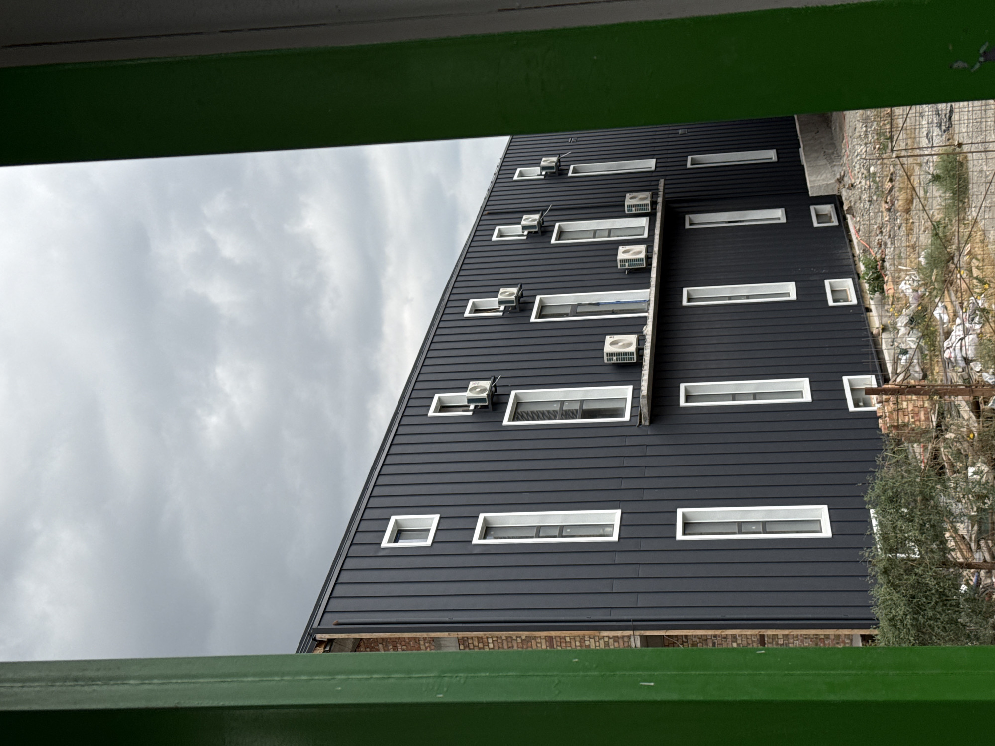 A medium shot captures the exterior of a dark grey, modern building against a cloudy sky, viewed through a vibrant green frame in the foreground. The building features a dark, vertical metal siding with numerous white-framed windows. Some windows are tall and narrow, while others are smaller and square. Several outdoor air conditioning units are mounted on the exterior walls, positioned above some of the windows. To the left of the building, exposed brickwork is visible, indicating construction or an unfinished section. The ground in front of the building is uneven and appears to be a construction site, with scattered debris, bricks, and fencing. The overall impression is one of an urban or industrial environment undergoing development.