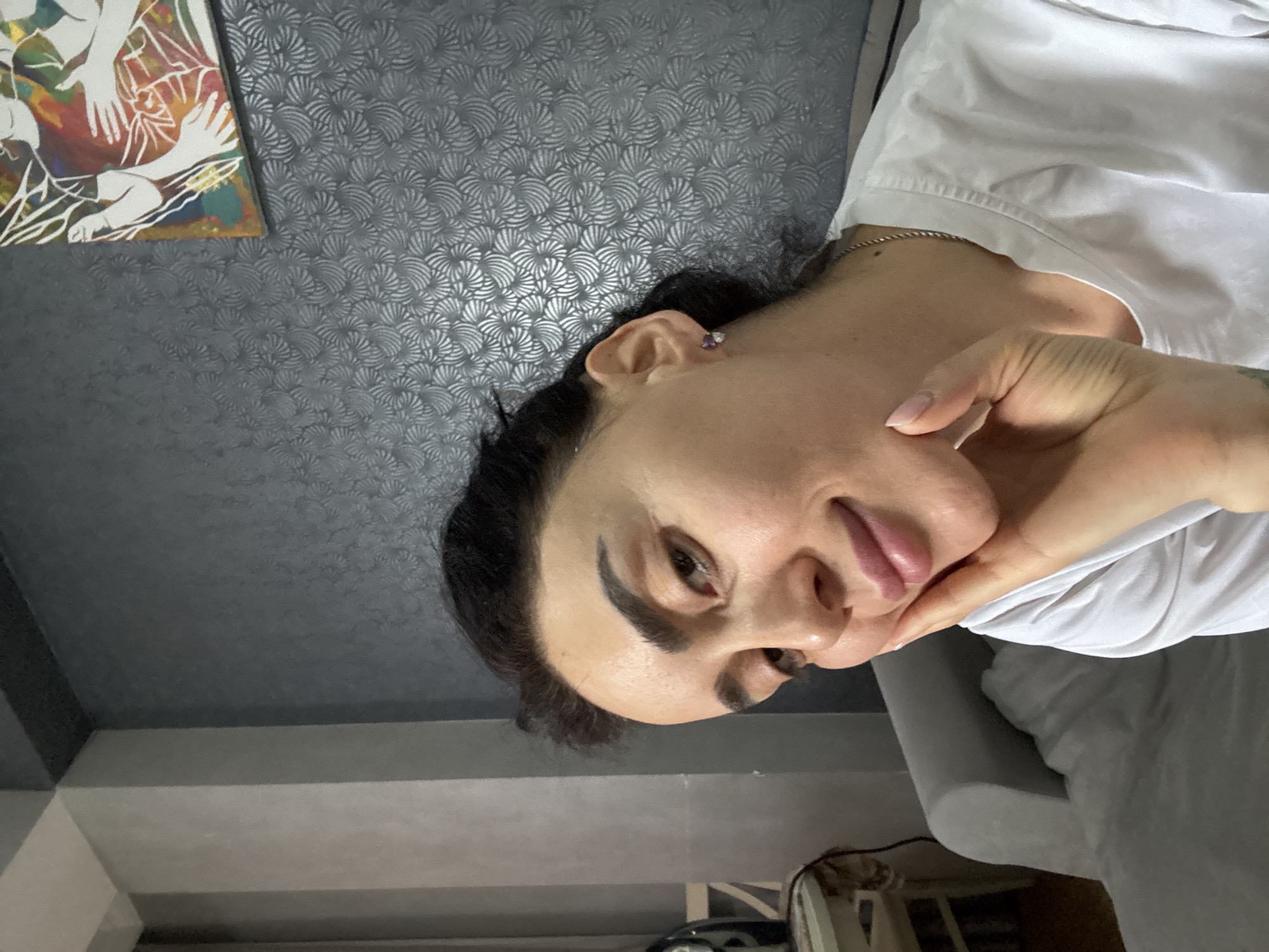 A woman with dark hair and fair skin, wearing a white shirt and a small earring, is looking directly at the camera. She is resting her chin on her hand. A painting is visible in the upper left corner of the frame. The background features a patterned wall. The location appears to be indoors, potentially in an apartment in Tbilisi, Georgia, based on the interior design. A gray couch is also visible.