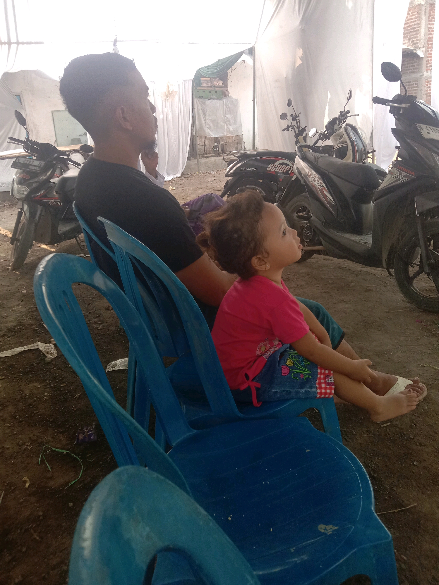 The image shows a man and a young girl sitting on blue plastic chairs, facing right, with several motorcycles parked behind them. The man, on the left, is wearing a black t-shirt and appears to be looking in the same direction as the girl. The girl, on the right, is wearing a pink t-shirt and a denim skirt with a checkered hem and floral embroidery. She is looking upwards and to the right. The background consists of white tents or tarpaulins, suggesting an outdoor event or temporary shelter. The ground is dirt and debris. The scene appears to be set in a somewhat unkempt outdoor area, possibly in a village or an area undergoing development, with brick structures visible in the far background. The presence of multiple scooters suggests a mode of transportation common in Indonesian urban or semi-urban environments. The overall lighting suggests it is daytime, possibly under bright, diffused light due to the white coverings. The context of Karang Anyar, Indonesia, a regency in Central Java, is consistent with the presence of these elements.