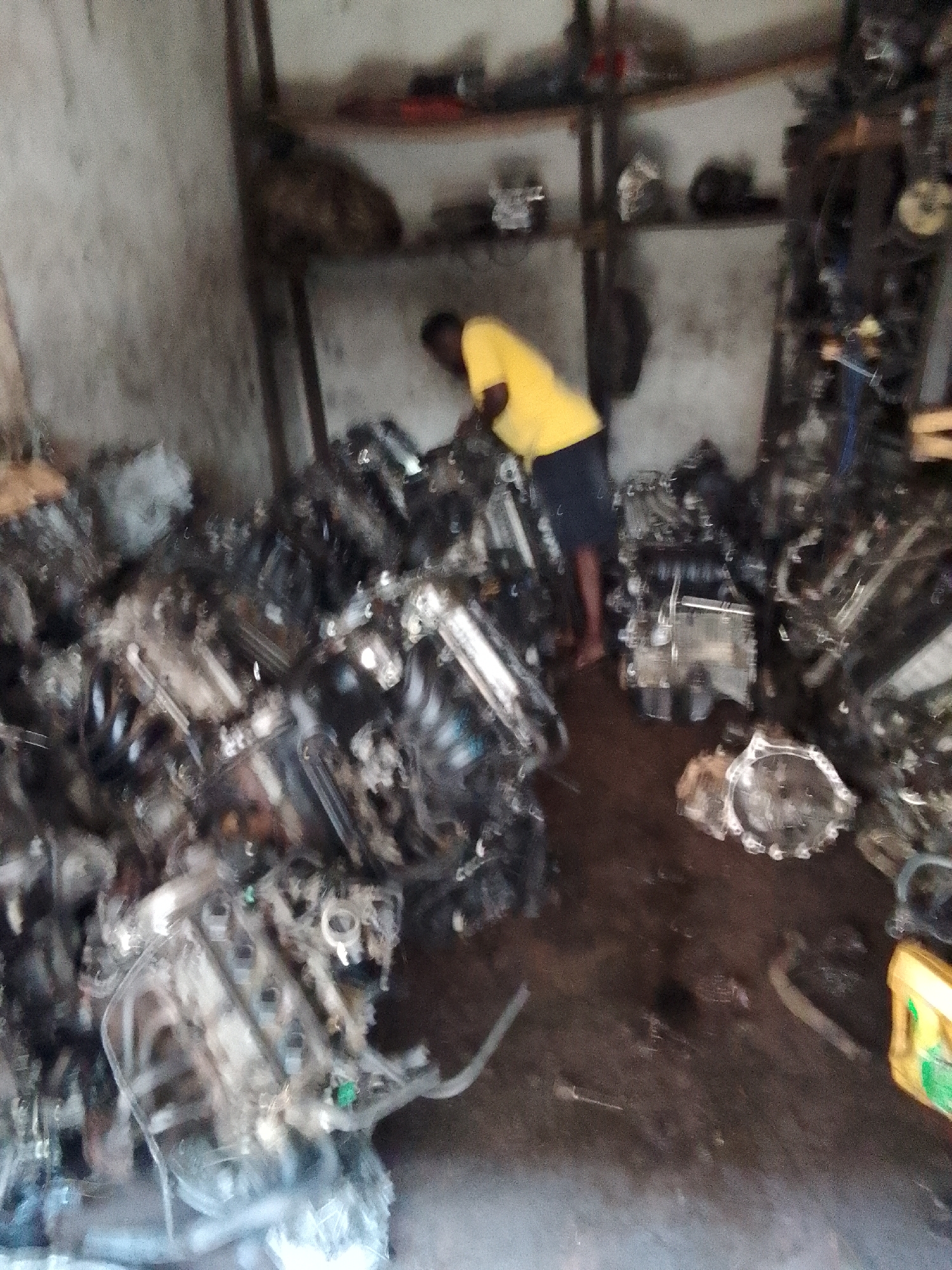 This image depicts an indoor scene, likely a workshop or storage facility for salvaged automobile parts in Onitsha, Nigeria. The central focus is a person, appearing to be male, wearing a bright yellow t-shirt and dark shorts, with bare feet. They are bent over, seemingly engaged in inspecting or sorting through a large assortment of car parts.
The setting is characterized by a cluttered environment, with numerous used car engines and various metallic auto components piled extensively across the dark, possibly dirt or concrete, floor. Several engine blocks, transmissions, and other mechanical parts are visible, suggesting a collection of items for sale or repair. In the background, wooden shelves line a light-colored wall, holding more auto parts and miscellaneous objects, further emphasizing the nature of the space as an auto salvage or repair shop. A yellow plastic container is visible on the floor in the lower right.
The activity taking place involves the individual working amidst these parts, suggesting tasks related to inventory, inspection, or dismantling. The lighting is diffused, indicating an indoor setting without direct sunlight. There is no discernible text visible anywhere in the image. The scene portrays a functional and industrious atmosphere, typical of a busy auto parts business.