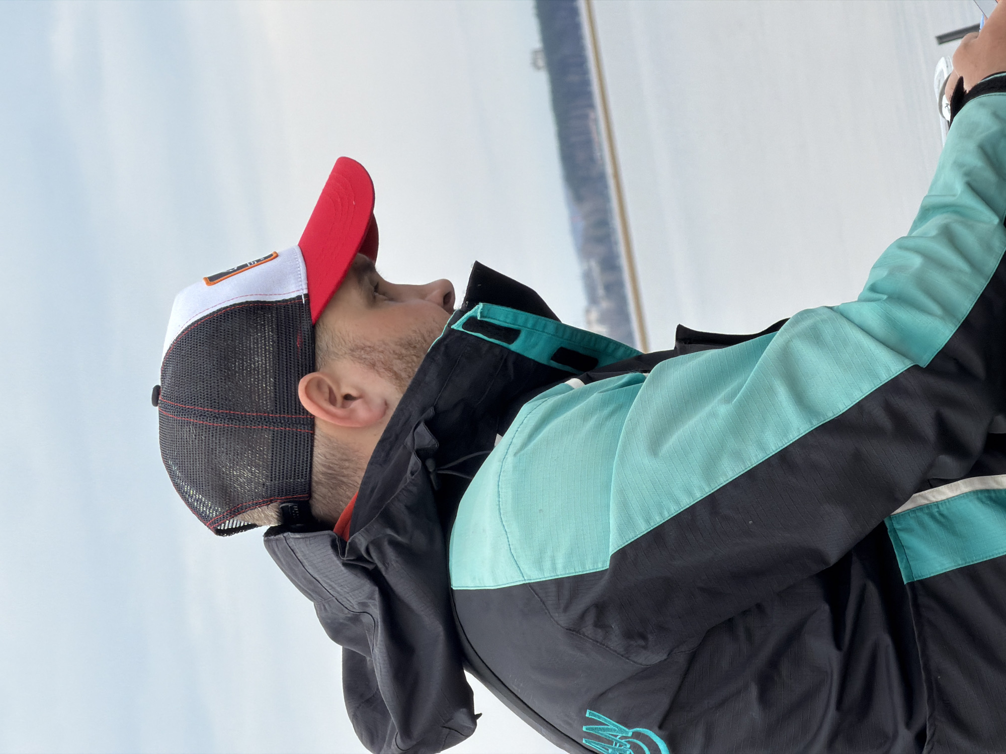 The image features a man from the chest up, oriented to the left. He is wearing a multi-colored baseball cap, with a red bill, white front, and black mesh back. He has a beard. He is wearing a black and teal jacket. The background appears to be a lake and a pale sky. Considering the location, it is likely the man is near or on the Votkinsk Reservoir near Chaykovsky, Russia.
