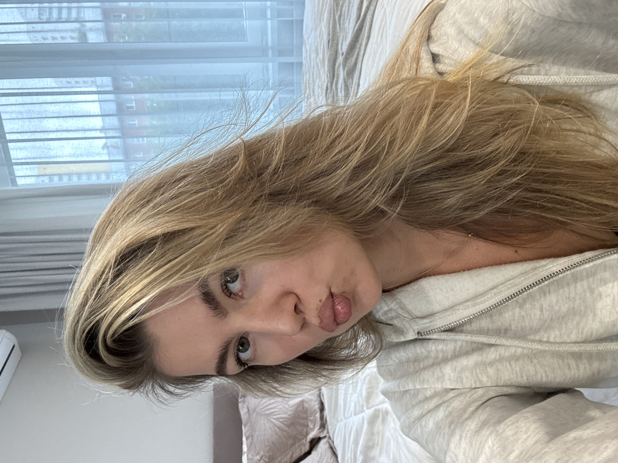In an indoor setting, a young Caucasian woman with blonde hair pulled back from her face and green eyes is captured in a close-up, head-and-shoulders shot. She is wearing a light gray zippered hoodie and a delicate gold necklace. Her lips are pursed in a kissing gesture, and she looks directly at the camera. The background reveals a bed with textured pillows and a white duvet, suggesting a bedroom environment. A window with sheer curtains and a glimpse of buildings outside indicates an urban location. An air conditioning unit is visible on the upper left wall. The overall lighting is soft and natural.
