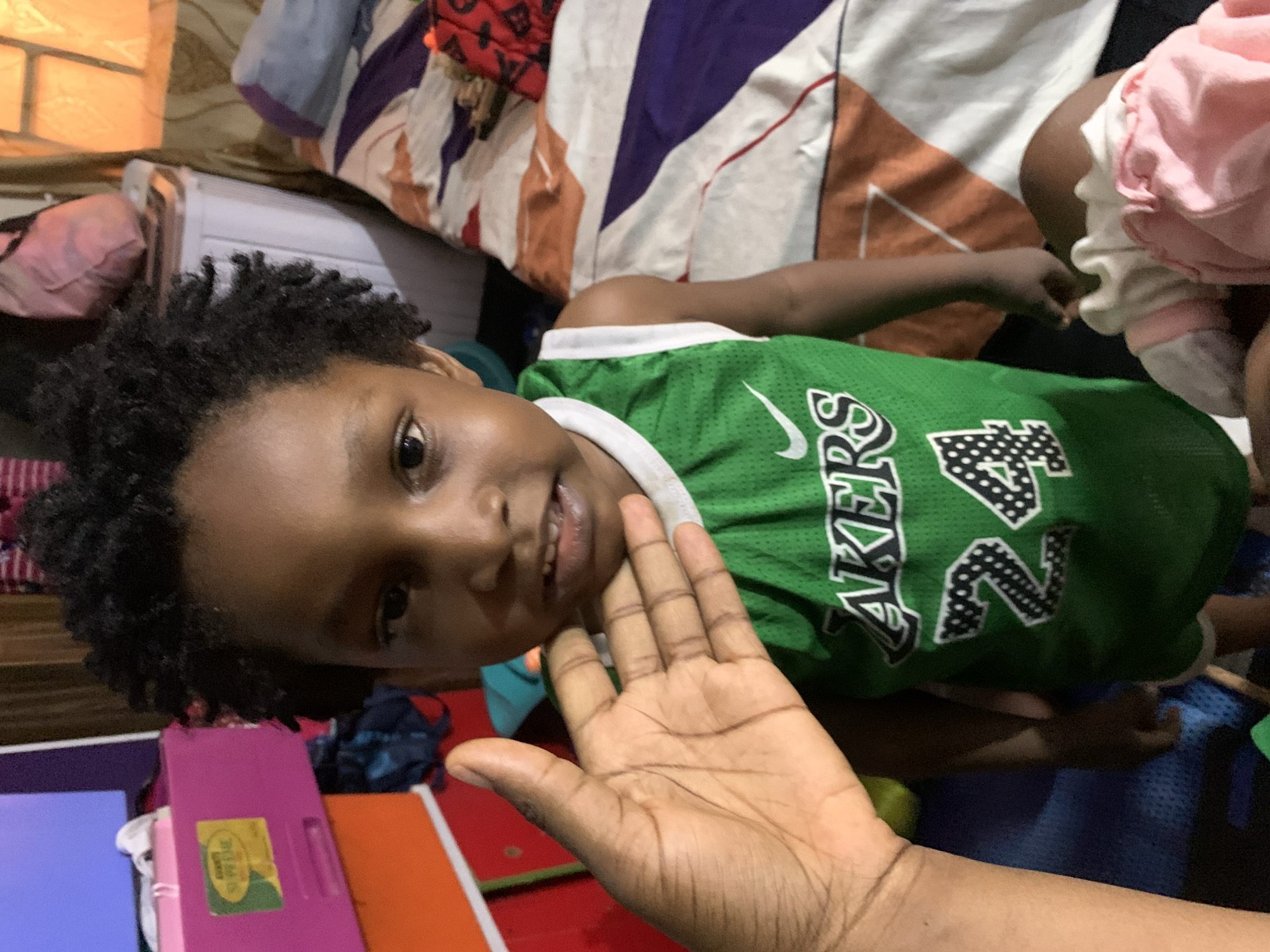 A child wearing a green and white Lakers jersey is lying on a patterned bed. A hand is raised near the child's face. The location is Rafin Bauna, Nigeria. Various items are visible in the background, including a pink container and fabric materials.