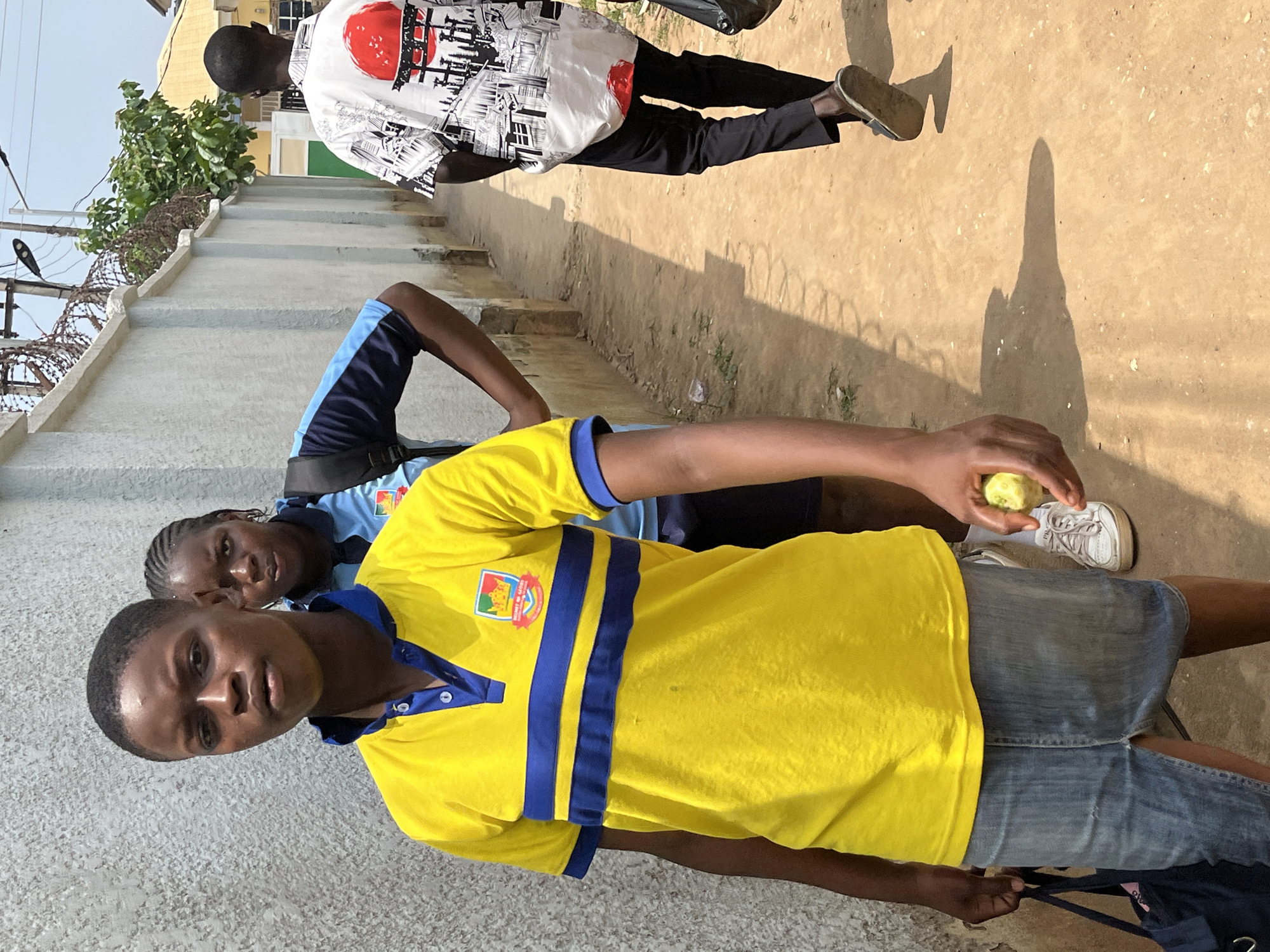 In Abuja, Nigeria, a group of individuals walk along a dusty, narrow path bordered by a grey concrete wall on the left and a lighter-colored wall on the right. In the foreground, a person wearing a bright yellow polo shirt with blue stripes and a denim skirt is facing the camera, holding a small, light-colored fruit in their right hand. They are also carrying a dark blue bag. Behind this person, another individual in a blue and black shirt with a backpack is partially visible. Further down the path, a person in a white shirt with a black and red Japanese-inspired graphic print walks away from the camera, dressed in black pants and sandals. The environment appears to be an urban or semi-urban setting, with a few trees and structures visible in the background. The ground is composed of loose, sandy dirt, and shadows are cast by the people and the surrounding structures.