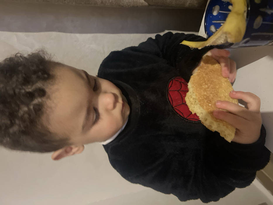 A child with curly hair, wearing a black fuzzy shirt with a red spiderweb design, holds a piece of toasted bread in their hands. The child's eyes are closed. A blue container with a yellow item, possibly a banana, is partially visible in the top right corner of the image. The background appears to be a couch.