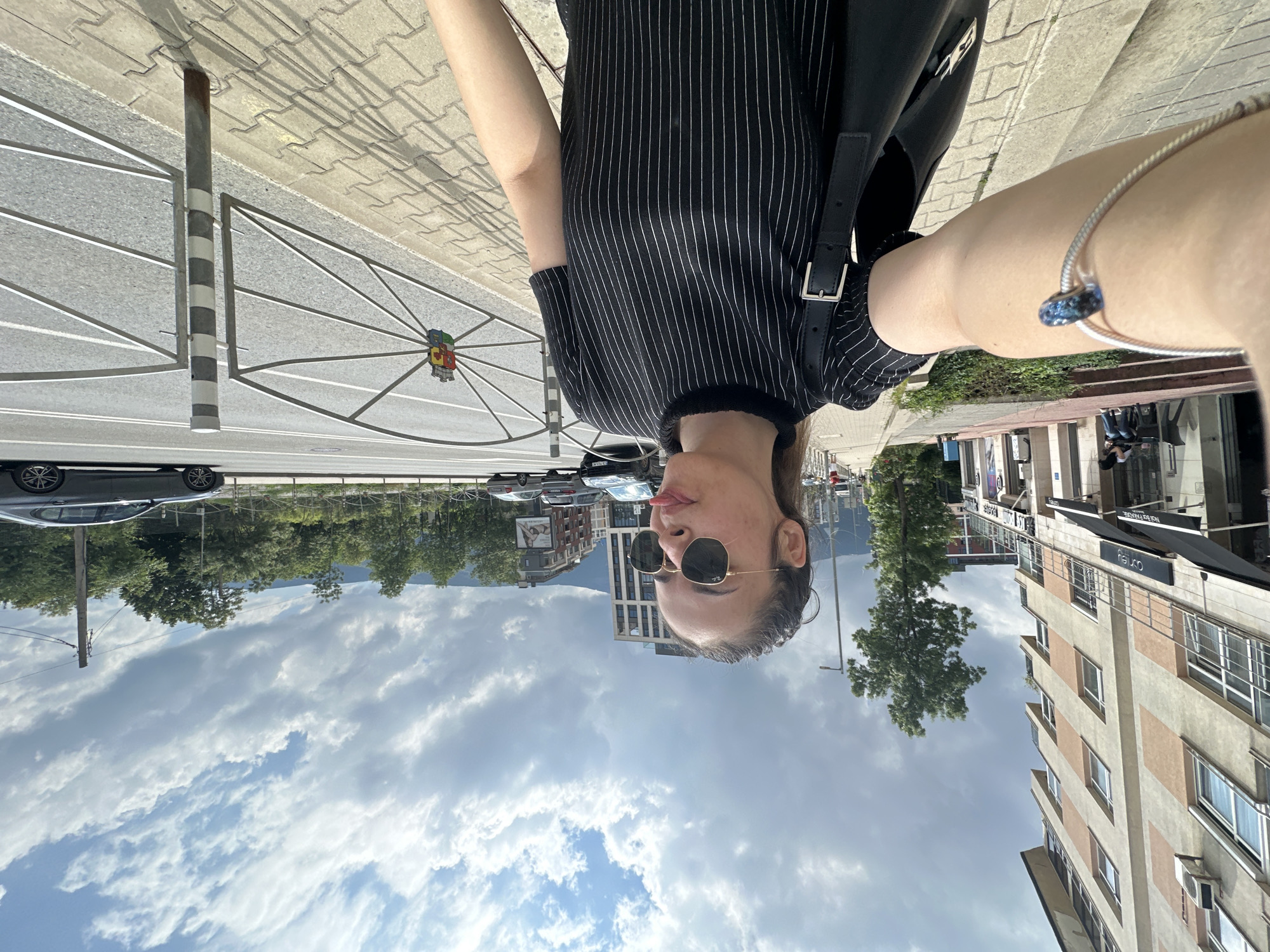 This media file captures a selfie taken outdoors in Sofia, Bulgaria, on a bright, sunny day. The primary subject is a person, likely female, seen from the chest up, wearing a black short-sleeved top with thin white vertical stripes and dark sunglasses. Her hair is pulled back, and she has a neutral expression, with her lips slightly parted. A silver-colored bracelet with a distinctive blue, possibly crystal or glass, charm is visible on her left wrist.
The background reveals an urban environment. To the left of the subject, a paved sidewalk gives way to a street where several cars are parked. A black and white striped pole stands prominently near a decorative metal gate. This gate features a colorful, shield-like emblem resembling a coat of arms, which could be a local municipal symbol. Further in the background, a line of green trees and taller buildings are visible, suggesting a city landscape with some natural elements. The sky above is largely clear with scattered white clouds. To the right of the subject, multi-story buildings line the street, some with visible commercial establishments on their ground floors. Partially visible text on these signs includes "HCU" or "HOU", "HOSPITALITY HOUSE", and a fragmented "S...Y". The scene depicts a lively city setting under clear weather conditions.