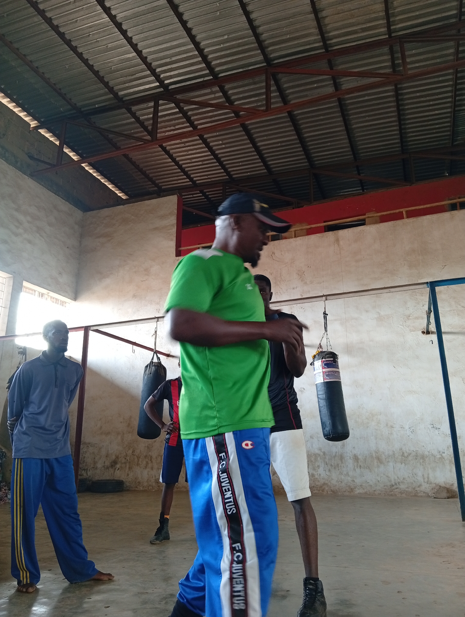 The image shows an indoor boxing gym with a corrugated metal roof and exposed beams. Three men are present. One man in the foreground, wearing a green t-shirt and blue track pants with "F.C. JUVENTUS" printed on the sides, is facing away from the camera and appears to be talking or instructing. To his left, a man in a gray long-sleeved shirt and blue track pants with yellow stripes is looking towards the foreground man. Further back, another man in a black and red shirt and white shorts is standing with his hands on his hips, looking towards the foreground. Two punching bags hang from the ceiling, one to the right of the man in black and red, and another further back and to the left. The scene appears to be during the daytime, as indicated by the light coming from windows on the left side of the image. The overall atmosphere suggests a training session or a casual gathering at a gym. There is visible text on the punching bag and on the track pants of the man in the foreground.