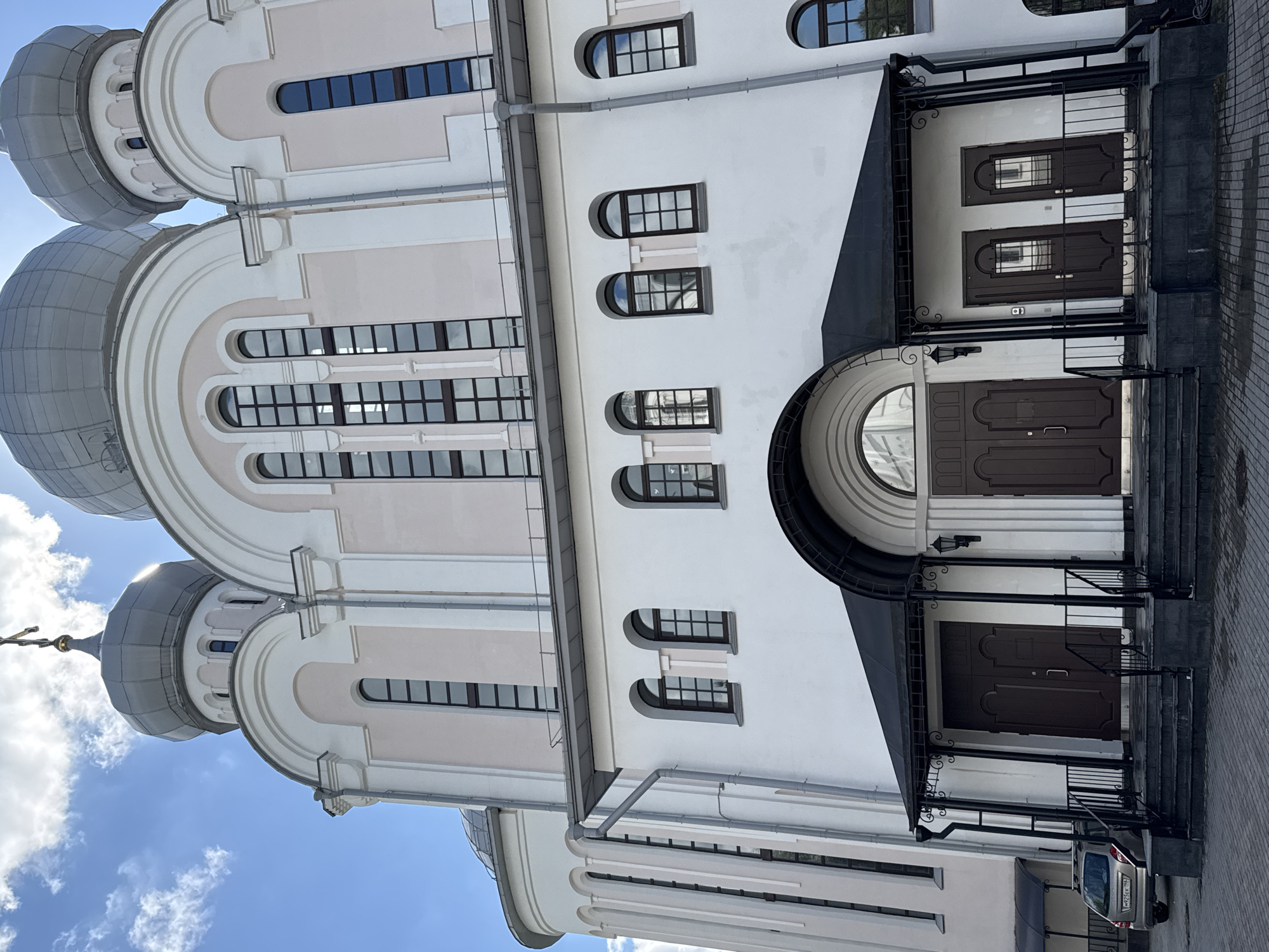 A low-angle, medium shot captures the facade of a white and light pink church building with gray domes against a bright blue sky with scattered white clouds. The building features multiple arched windows with dark frames and grids, as well as tall, narrow windows on its upper sections. A dark gray, sloped roof extends over the entrance, supported by black metal posts with decorative scrollwork. Two large, dark brown double doors are set within the entrance, accessible by a set of dark gray stairs and a black metal railing. To the left of the main entrance, a silver hatchback car is parked on a paved area. The architecture suggests a religious building, possibly an Orthodox church, given the onion-shaped domes and cross visible on the topmost dome. The scene appears to be a daytime shot in an urban setting, likely Saint Petersburg, Russia, given the architectural style and prevalence of Orthodox churches in the city. No people are visible in the frame, and the car is stationary.