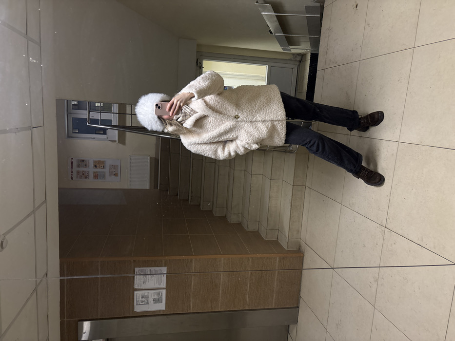A single individual stands in a common area of a residential building in Kudrovo, Russia, capturing a mirror selfie. The subject, identifiable by visible long blonde hair, is dressed in a large white faux-fur hat, a light beige textured coat with a prominent dark button closure, dark-colored trousers cuffed at the ankles, and dark brown lace-up boots. Both hands hold a light pink smartphone directly in front of the face, obstructing the view of facial features. The immediate environment includes a light-tiled floor reflecting overhead lighting. To the left, a large vertical mirror, used for the selfie, reflects an elevator with a metallic or wood-paneled finish and two framed notices on the adjacent wall. To the right, a staircase with light-colored steps ascends, accompanied by a metal handrail. Above the staircase, a white radiator is affixed to the wall, below an information board displaying multiple documents or posters. A light-colored door with a small rectangular window is visible in the background to the far right.