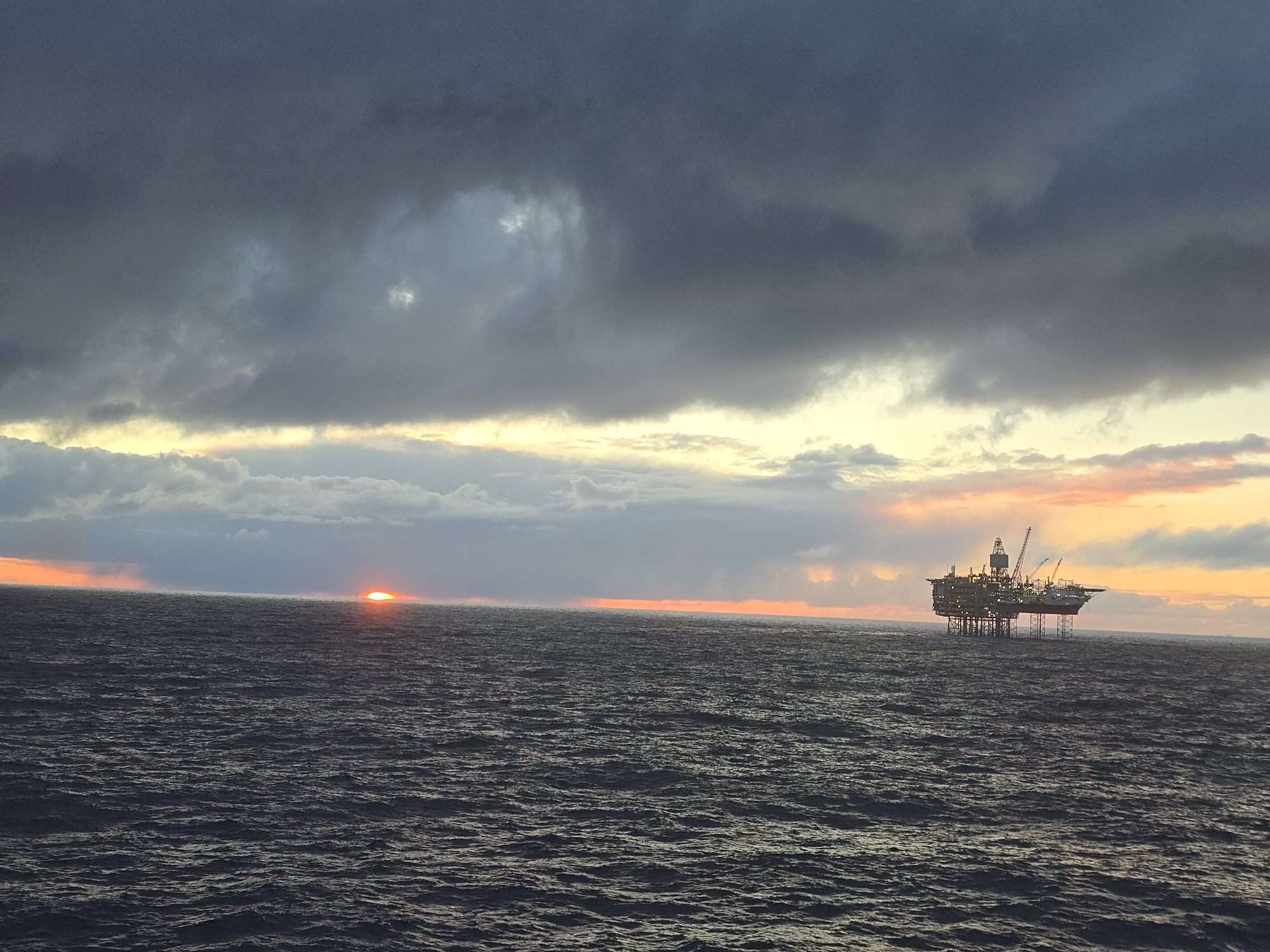 The image shows an offshore oil platform in the ocean at sunset. The sky is filled with dark, heavy clouds, but there is a break in the clouds on the horizon where the sun is setting, creating a bright orange glow on the water. The oil platform is located some distance from the camera and is silhouetted against the setting sun. The ocean is choppy, with visible waves. The location is unknown.