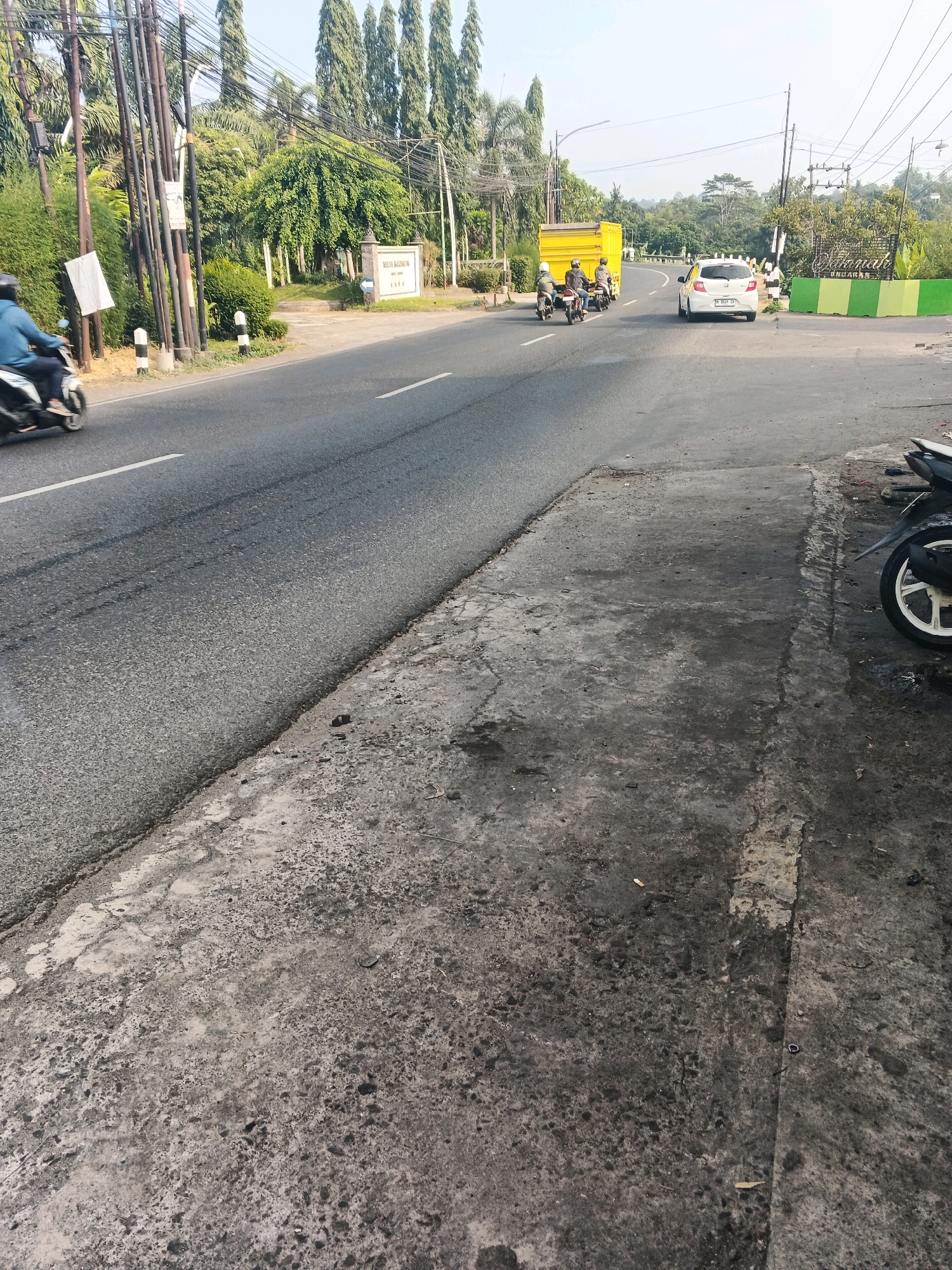 A bright sunny day shows a road in Ungaran, Indonesia, where a yellow truck is turning left, followed by three motorcycles. A white car is in the right lane of the road. On the left side of the road is a sidewalk and a grassy area with trees. On the right side of the road is a parked motorcycle on the sidewalk.