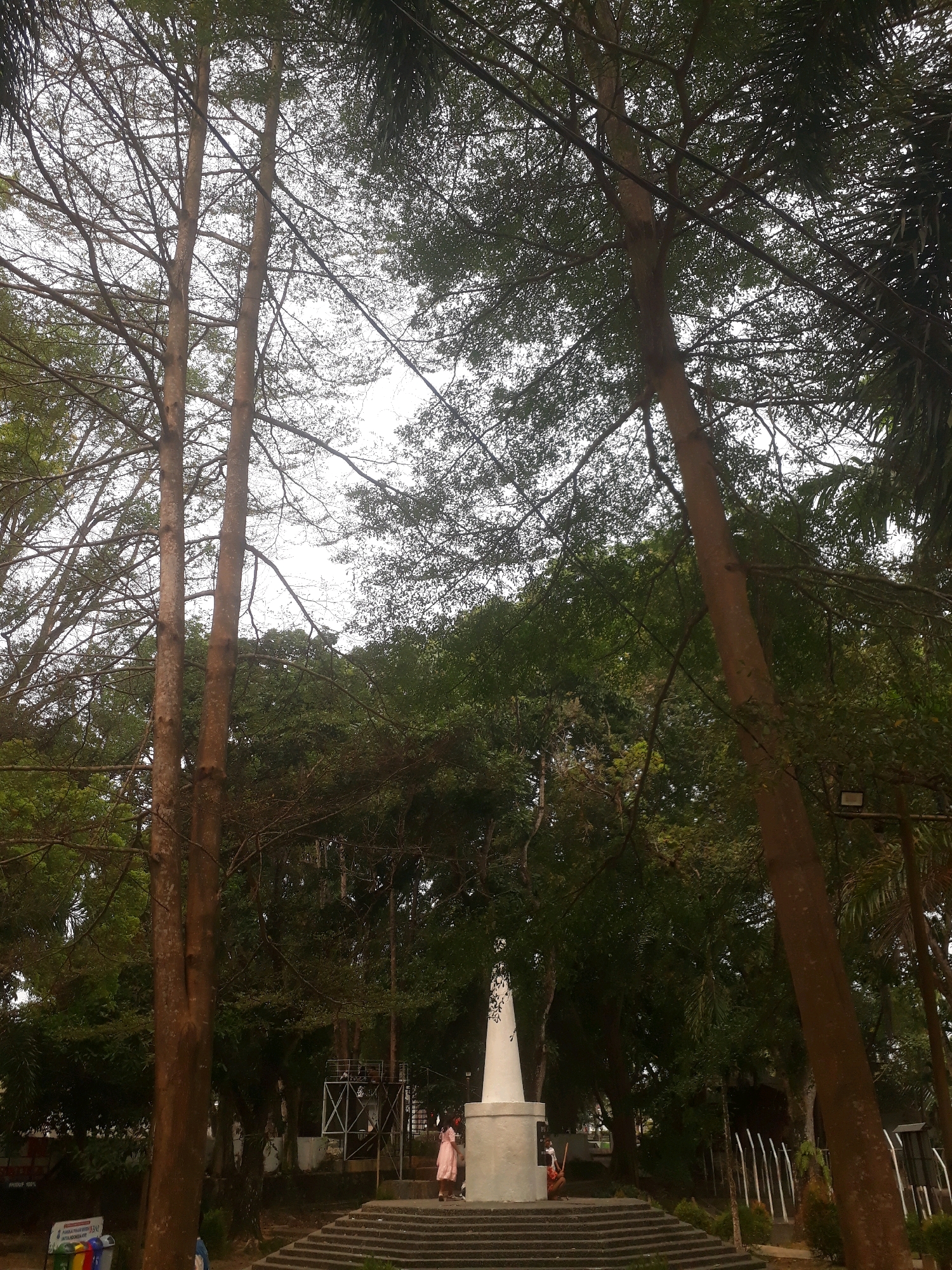In Pangkalpinang, Indonesia, the image captures a monument at the heart of a park-like setting. The monument, a tall, white, conical structure, stands on a stepped base surrounded by lush greenery. Two individuals are present near the monument; one, wearing a pink dress, stands on the steps, while another, partially visible, is positioned behind the monument. The scene is framed by the dense foliage of trees, with their branches creating a natural canopy overhead. The sky above is overcast, casting a diffused light on the environment. To the left, a white sign with text and colorful bins are visible at the base of the monument's steps, suggesting a public space.