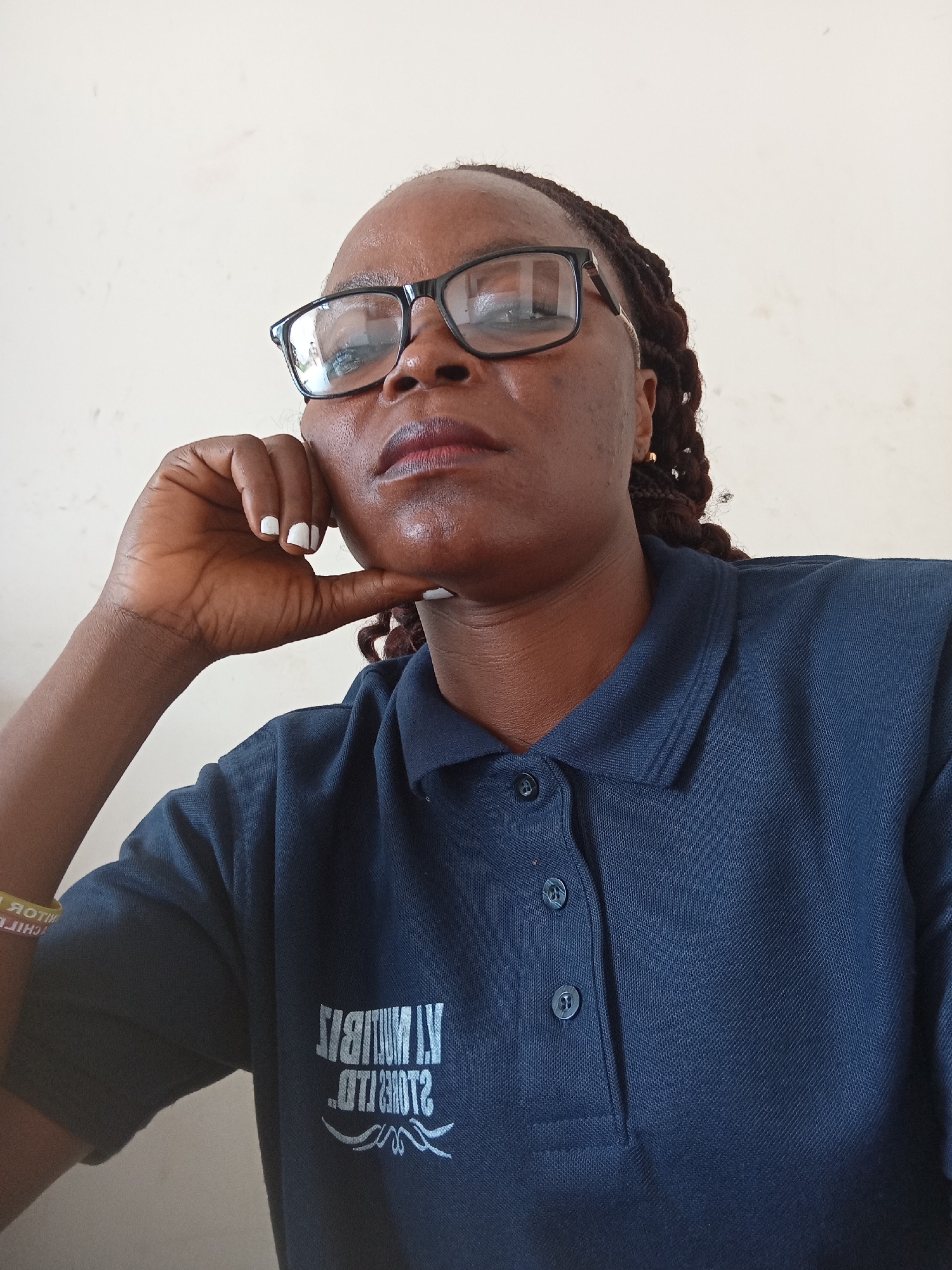 This is a close-up, eye-level portrait of a Black woman wearing glasses and a dark blue polo shirt. She is resting her chin on her hand, with her elbow propped up. Her expression is thoughtful or serious, with her gaze directed upwards and slightly to the left. She has dark, braided hair and her fingernails are painted white.
The polo shirt has a collar and two buttons visible at the neckline, one of which is slightly ajar. There is white text and a graphic design on the left side of the shirt, with the text appearing to be a logo. A yellow bracelet with red text is visible on her left wrist, and the text appears to be in reverse, suggesting it might be a reflective surface or seen through the camera.
The background is a plain, light-colored wall. The lighting suggests an indoor setting, possibly during the daytime. The overall mood is contemplative.
Visible text on the shirt appears to read "ALMUBIT" and "IT'S MORE THAN". The text on the yellow bracelet is difficult to decipher clearly but appears to include "CHIF".