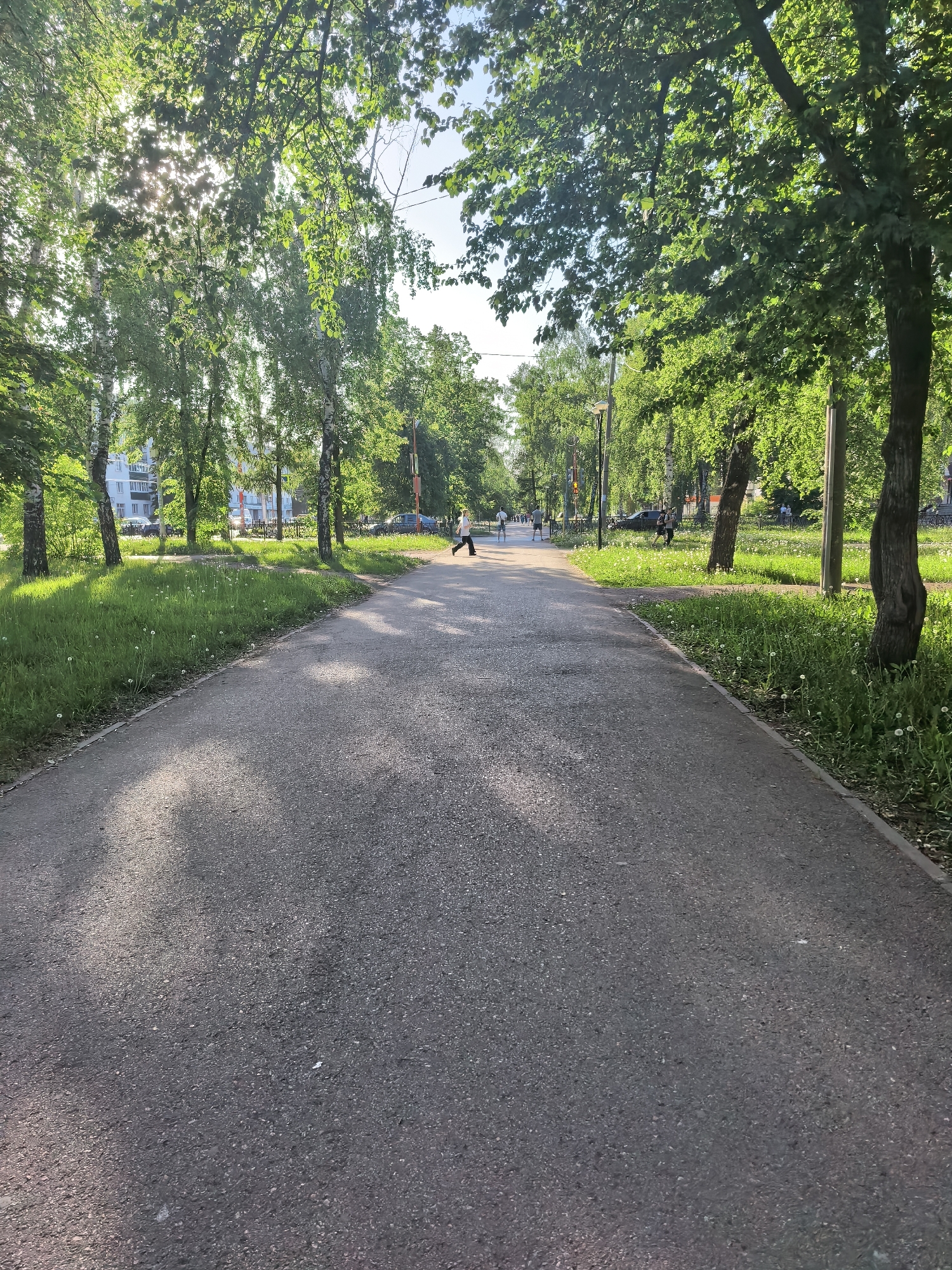 A paved walking path stretches into the distance, flanked by green grass and trees. A person is seen jogging on the path, with other people visible further away. Sunlight filters through the leaves of the trees. The location is a park area in Sterlitamak, Russia. The environment is a sunny day.