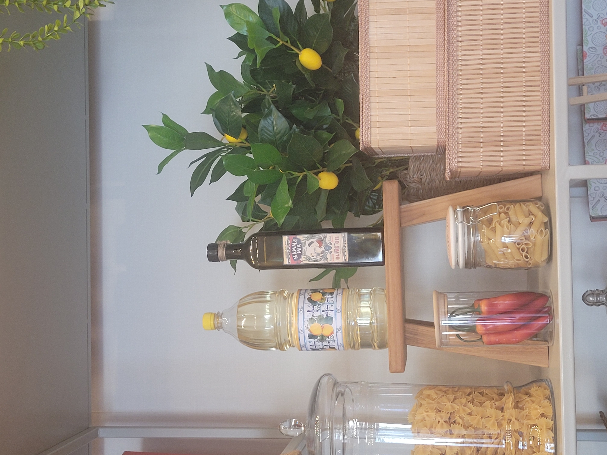 The image depicts a well-organized kitchen shelf display in Limassol, Cyprus. The foreground is dominated by a large glass jar filled with farfalle pasta on the left, and a smaller glass jar containing penne pasta on the right. Between these, a clear glass cylinder holds three red chili peppers. A wooden stool supports two bottles: a large plastic bottle of cooking oil with a decorative label featuring lemons, and a dark glass bottle of "Mamma Mia Olive Oil" with a traditional label. Behind the bottles, a vibrant green plant with small yellow fruits, possibly a citrus tree, is displayed. Two woven bamboo baskets, one stacked on top of the other, are positioned to the right of the plant. The background consists of a plain, light-colored wall and a white shelf. The lighting is soft and diffused, suggesting an indoor setting. There are no people or animals present. The scene is static and suggests a retail or home display of food products and decorative items.