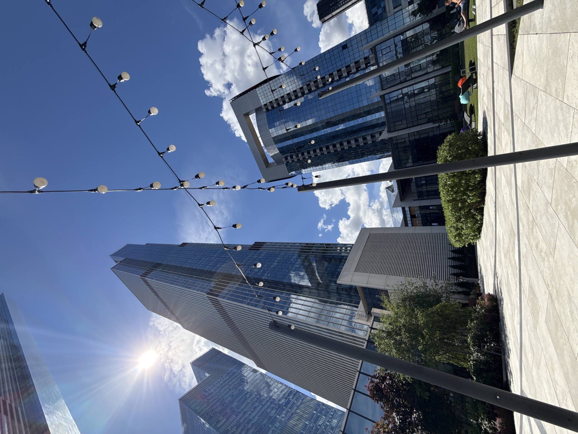 The image depicts a sunny day in Moscow City, Russia. The perspective is from a paved plaza looking up at several modern skyscrapers with glass facades that reflect the blue sky and white clouds. A string of decorative lights hangs across the foreground. In the middle ground, there are landscaped areas with trees and bushes, as well as lounge chairs on a grassy area where two individuals are visible, one sitting and one lying down. The overall scene suggests a contemporary urban environment, possibly a business or residential complex with public outdoor spaces.