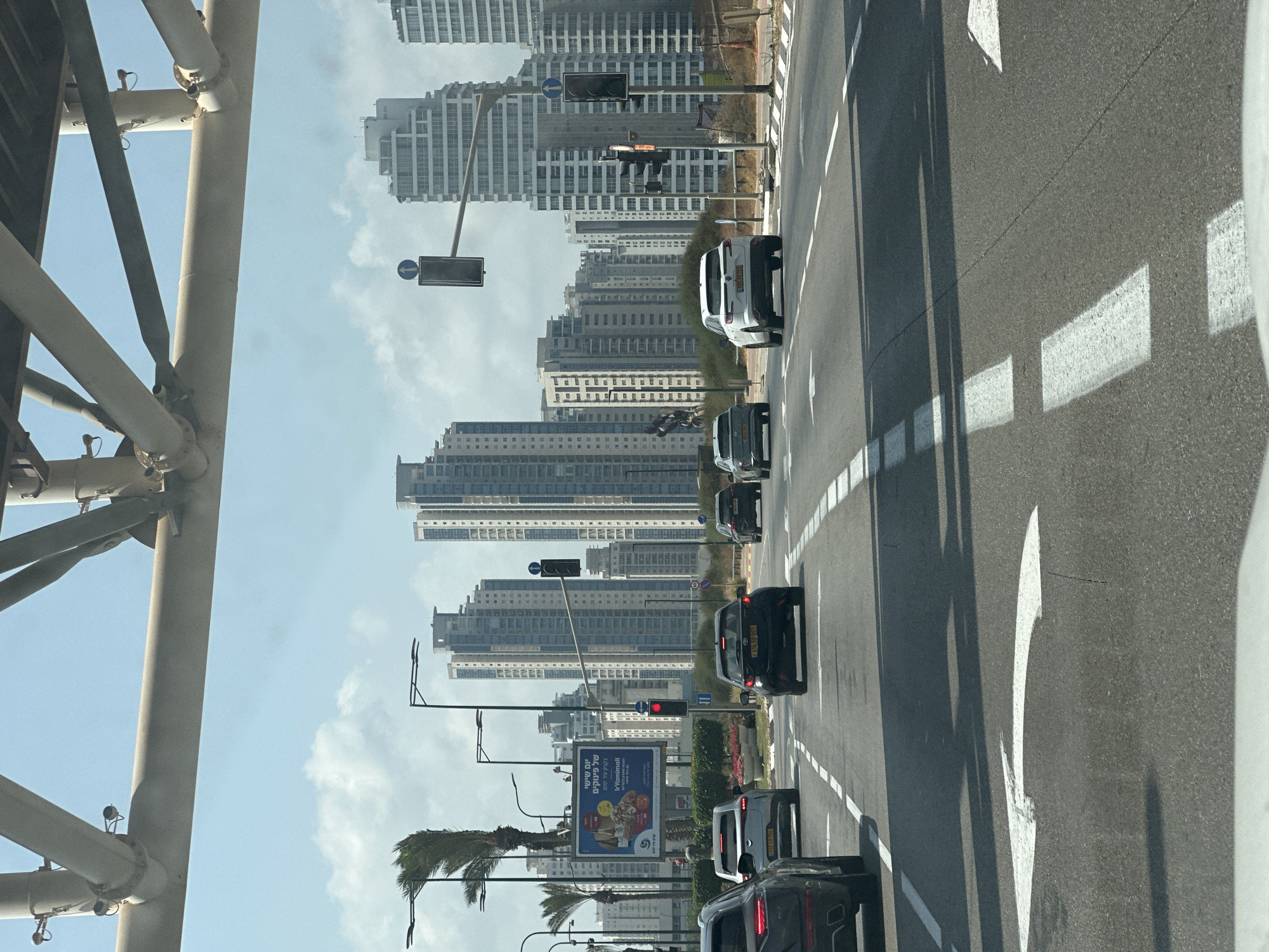 The image depicts a street scene in Netanya, Israel, with a focus on traffic and urban architecture. In the foreground, a road with white lane markings is visible, with several cars driving on it. One car in the left lane has a turn arrow painted on the road in front of it. In the background, a cluster of modern high-rise buildings dominates the skyline under a partly cloudy sky. Several traffic lights are visible, one of which is red, indicating a stop. A billboard is also present on the left side of the road, displaying text in Hebrew and English. Palm trees and other greenery are scattered throughout the scene, suggesting a relatively warm climate. The overall impression is one of a busy urban environment.