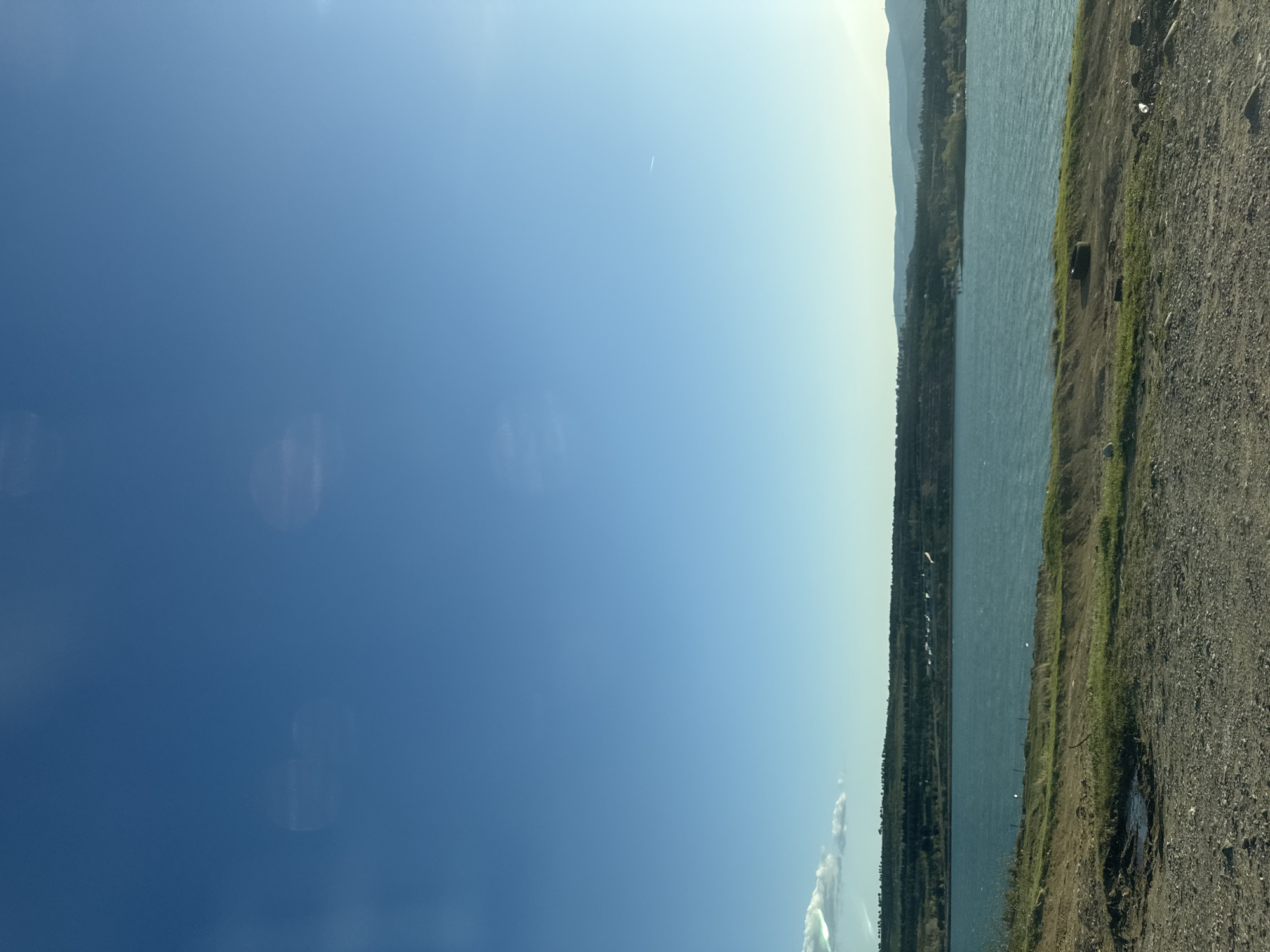 The image shows a lake bordered by a gravelly shoreline. On the far side of the lake, trees line the shore, with a distant mountain range visible in the background. The sky occupies a large portion of the image.
Without further information, determining the precise location in Tbilisi, Georgia is impossible. Additional details such as landmarks, specific vegetation types, or distinctive architectural features would be needed to narrow down the location.