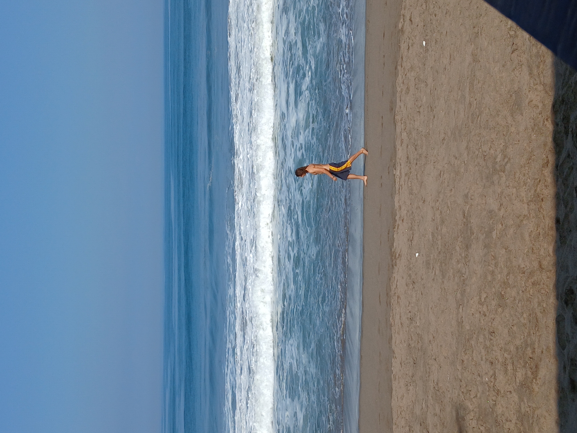 A full shot shows a lean, long-haired male with a tan walking on a beach in North Kuta, Indonesia. The man is wearing dark blue and yellow swim trunks. He is walking from left to right, facing to the left. The beach is sandy and there are footprints visible in the sand. The ocean is calm with small waves. The sky is clear and blue.