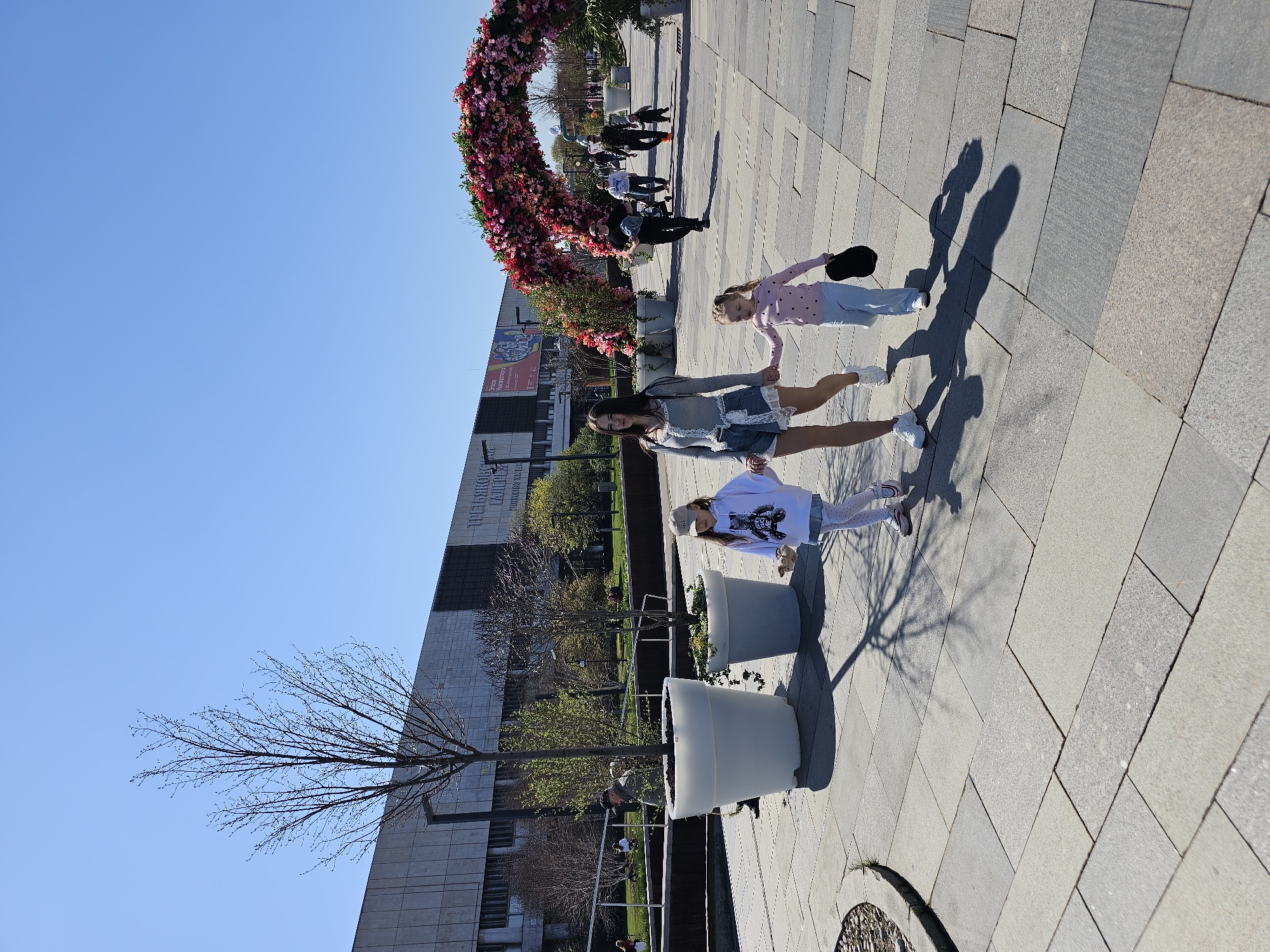 The image captures an outdoor scene in Moscow, Russia, featuring a mother and two daughters walking on a paved area. The mother is in the center, holding hands with her daughters on either side.
In the background, a large floral archway is visible, along with several other people walking in the distance. To the left, a tall building is identifiable as the Tretyakov Gallery, based on the partially visible signage in Cyrillic script. Adjacent to the path, there are large, cylindrical planters. The sky is clear and blue, suggesting a sunny day.