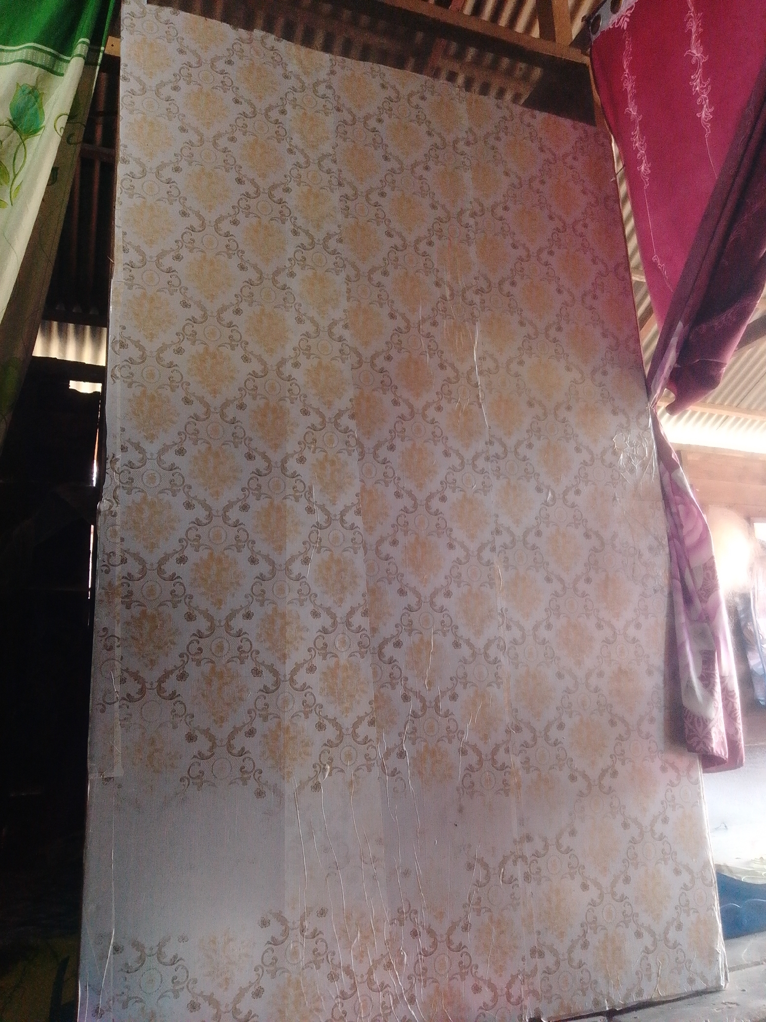 The image shows a large, flat object with a patterned surface, possibly a mattress or a piece of wall paneling, leaning against a wooden structure. The pattern is floral or damask-like, with beige and white tones. To the left, a green curtain with a floral design is visible. To the right, a pink curtain hangs. The background suggests an outdoor or semi-outdoor setting with visible wooden beams and corrugated roofing. Light streams in from the right, creating shadows and highlights. There are no people present in the image. The scene appears to be in a humble dwelling or storage area, consistent with a rural or semi-urban environment. The location of Labuhan Bilik, Indonesia, is provided as context, suggesting a tropical setting.