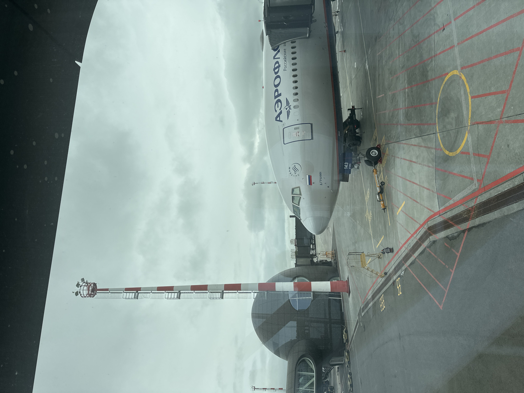 An Aeroflot passenger airplane is parked at a gate at an airport on a cloudy day. The airplane is silver with blue lettering on the side that reads "AEROFLOT" and "Российские авиа". It is connected to a jet bridge. To the left of the airplane is a large, dark, dome-shaped building with reflective panels. A tall, red and white striped tower with lights at the top is located between the airplane and the building. The ground is asphalt with yellow and red markings. The image appears to be taken from inside a terminal building, looking out a window. The scene depicts a typical airport environment.