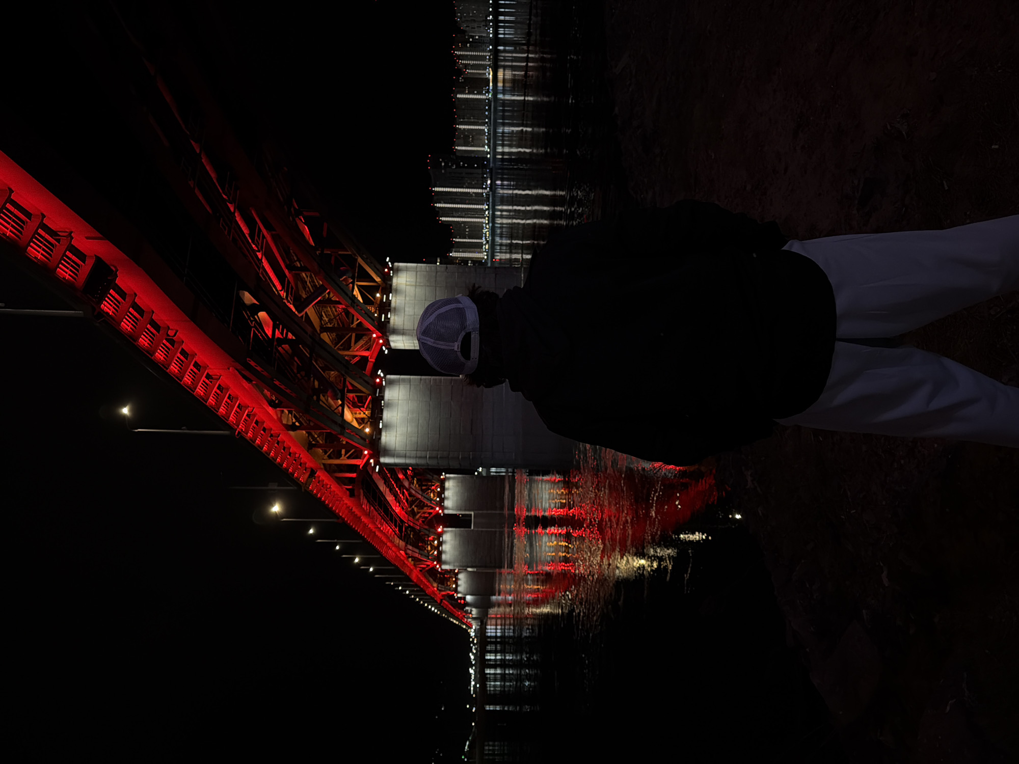 A person wearing a dark jacket, light-colored pants, and a gray baseball cap is standing with their back to the viewer. They are positioned on a dark, dirt surface, likely near a riverbank. In front of them is a bridge illuminated with red lights, which reflect in the water below. On the other side of the river, building lights shine, also reflecting in the water. The scene is dark, with most details obscured except for the illuminated bridge, water reflections, and the partially visible person. This scene is the Communal Bridge in the city of Krasnoyarsk, Russia.