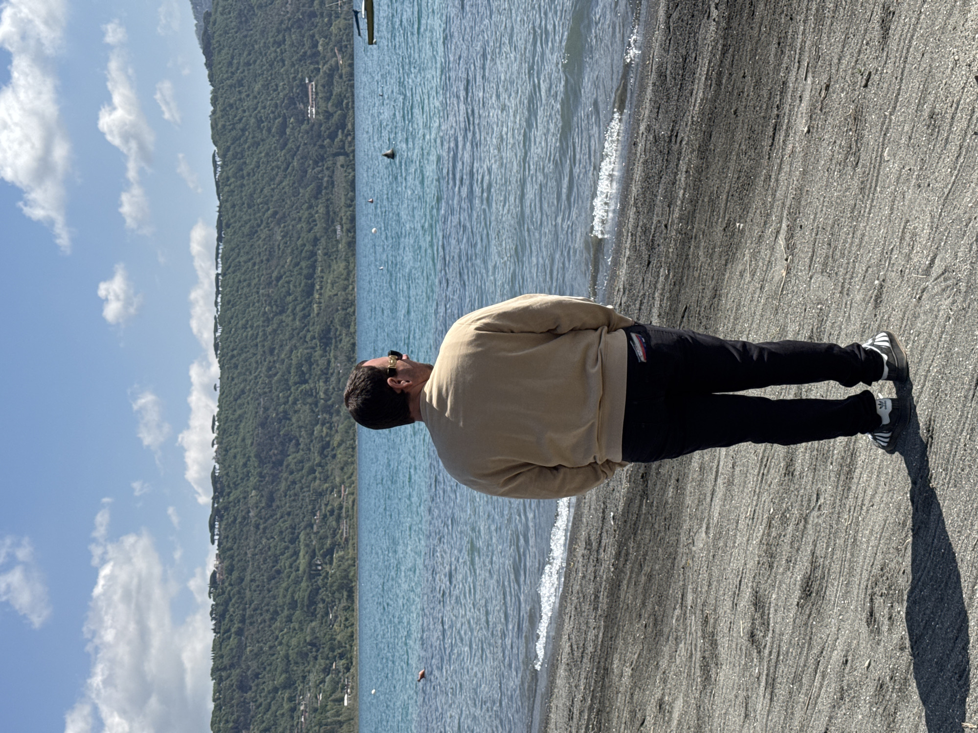 A person stands on a dark, pebbly shore, gazing out at a serene body of water, likely Lake Albano in Castel Gandolfo, Italy. The individual, seen from behind and slightly to the side, is a man with dark hair, wearing sunglasses on his head. He is dressed in a light tan or beige long-sleeved top and black trousers with a small, rectangular red, white, and blue patch visible on his left hip pocket. His feet are clad in dark sneakers with white stripes, resembling an Adidas design.
The tranquil lake stretches across the mid-ground, its surface showing gentle ripples and small waves lapping at the shore. Several small objects, possibly buoys or markers, are scattered across the water, including a distinct triangular black shape. Beyond the lake, a majestic, tree-covered mountain or large hill forms the backdrop, rising steeply and appearing lush green under a clear blue sky. Scattered white clouds drift overhead, indicating pleasant weather.
The scene conveys a sense of calm observation or contemplation, with the man seemingly lost in the expansive view of the water and the natural landscape. The time of day appears to be sunny, possibly late morning or early afternoon, with the sun casting a discernible shadow of the person on the pebbly ground to his right. There is no clearly legible text visible on clothing or objects, other than the distinctive three white stripes on his sneakers.