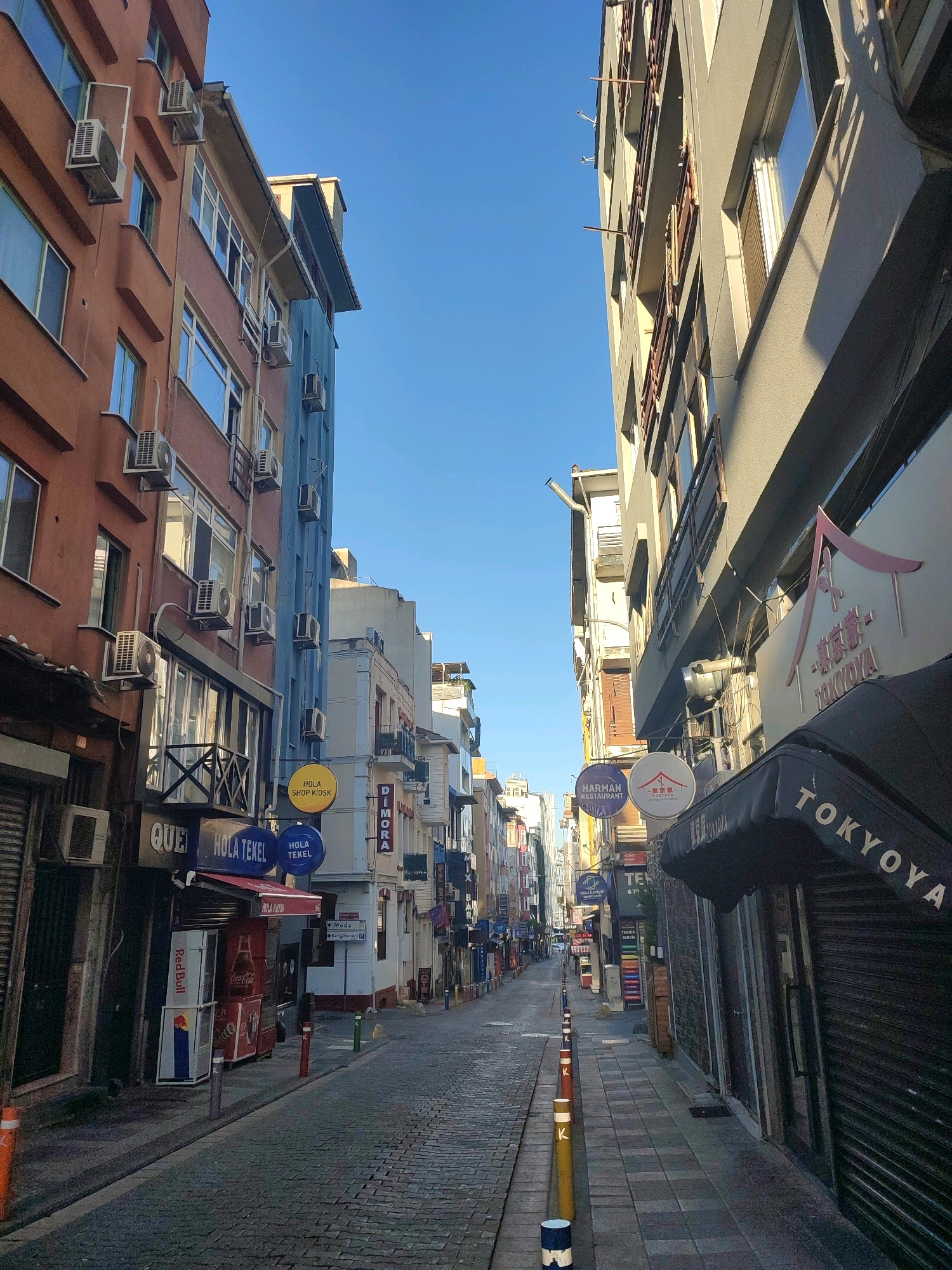 The image depicts a narrow, cobblestone street in Kadıköy, Turkey, lined with multi-story buildings. The buildings are a mix of colors, including orange, blue, and gray, and feature numerous windows and air conditioning units. Several businesses are visible along the street, indicated by signage such as "HOLA SHOP KIOSK," "DIMORA," "HARMAN RESTAURANT," and "TOKYOYA." Some storefronts have metal security shutters drawn down. The street is empty of people and vehicles, creating a quiet and still atmosphere. The sky is clear blue, suggesting a sunny day. The perspective is looking down the street, with the buildings creating a canyon-like effect.