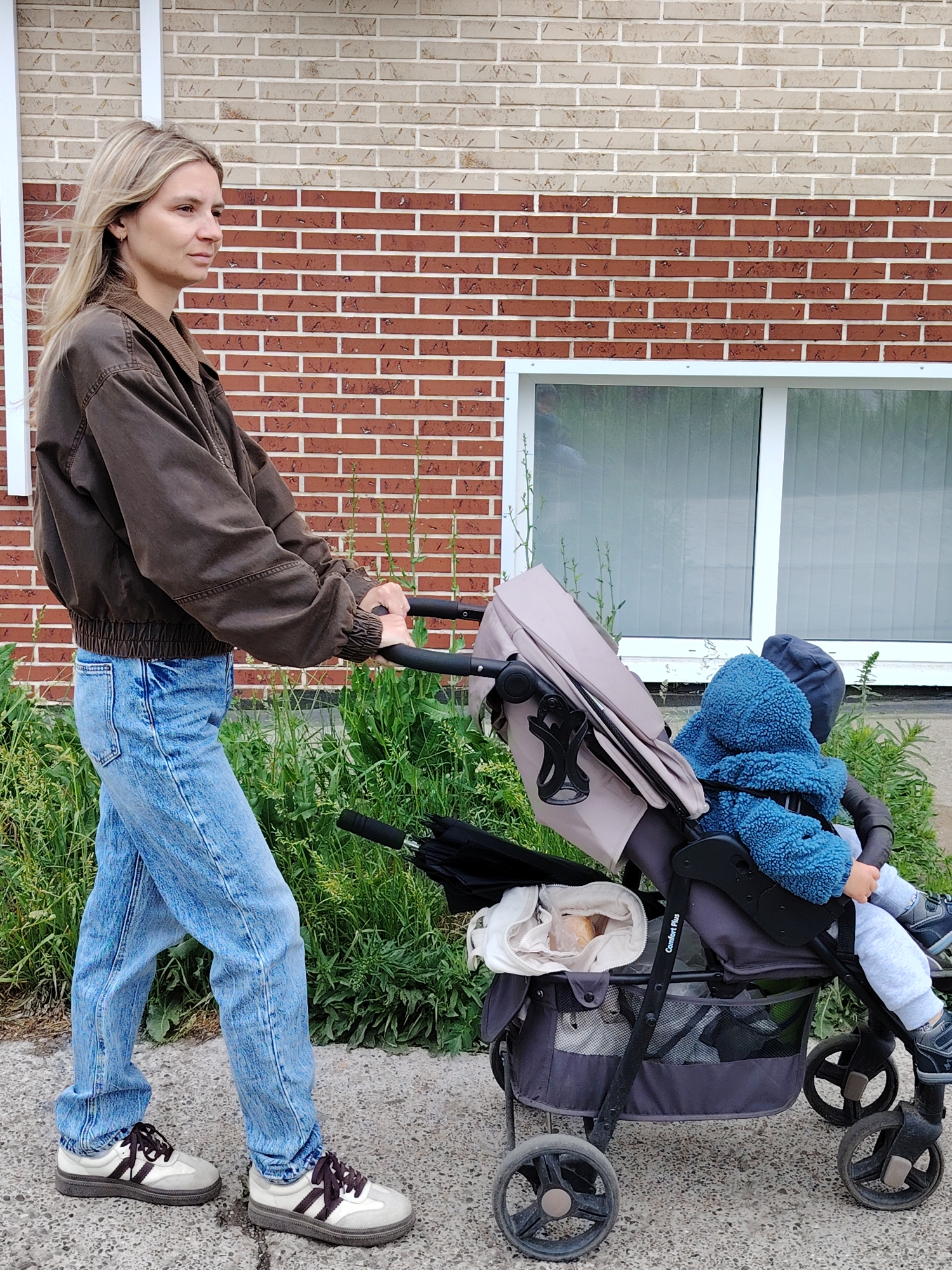 A woman pushes a baby stroller on a sidewalk in Chaikovsky, Russia. The woman, likely in her late 20s to early 30s, has light hair and is wearing a brown jacket, blue jeans, and white sneakers with brown laces. The baby stroller contains a child bundled in a blue hooded jacket and gray pants. A white tote bag, likely containing bread, is placed in the stroller's basket underneath the seat. The setting appears to be a residential area with a brick building behind the woman and a window. Green plants grow along the side of the building.