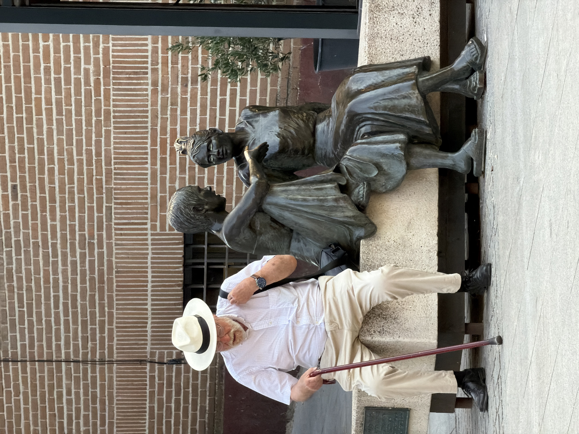 An elderly Caucasian man with a white hat, white short-sleeved shirt, khaki pants, black shoes, and glasses is sitting on a stone bench with a cane in his right hand. He is looking down. Next to him are two bronze statues of women, one facing him and appearing to be speaking, the other looking away. The statues are seated on the same bench. Behind the bench is a brick wall. To the right of the statues is a potted plant. This scene is located in the city of Las Rozas de Madrid, Spain.