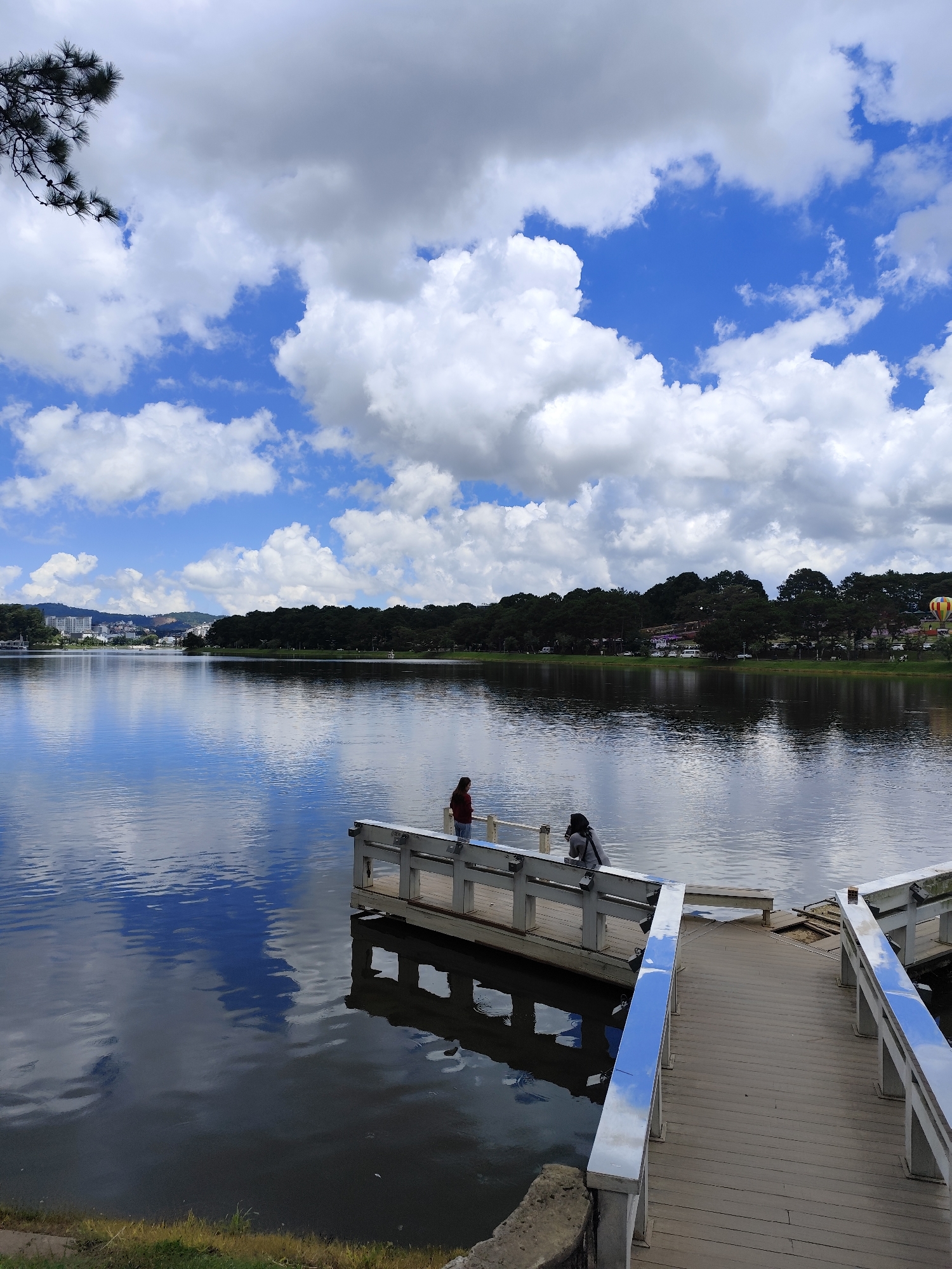 The image shows Xuan Huong Lake in Da Lat, Vietnam, on a cloudy but bright day. The lake's surface is calm and reflects the sky, clouds, and the surrounding treeline. In the foreground, a wooden pier extends into the water. Two individuals are visible on the pier. One, wearing a red top, stands near the edge of a section of the pier. Another, wearing a grey top, is positioned further down the pier, facing away from the camera. To the right, a pathway made of wooden planks leads from the foreground towards the lake, with white railings along its sides. The far side of the lake is lined with a dense forest, and beyond that, buildings and hills are visible, suggesting the presence of the city. A hot air balloon, yellow and red, is suspended in the sky in the distance. A pine tree branch with needles hangs from the top left corner of the frame.