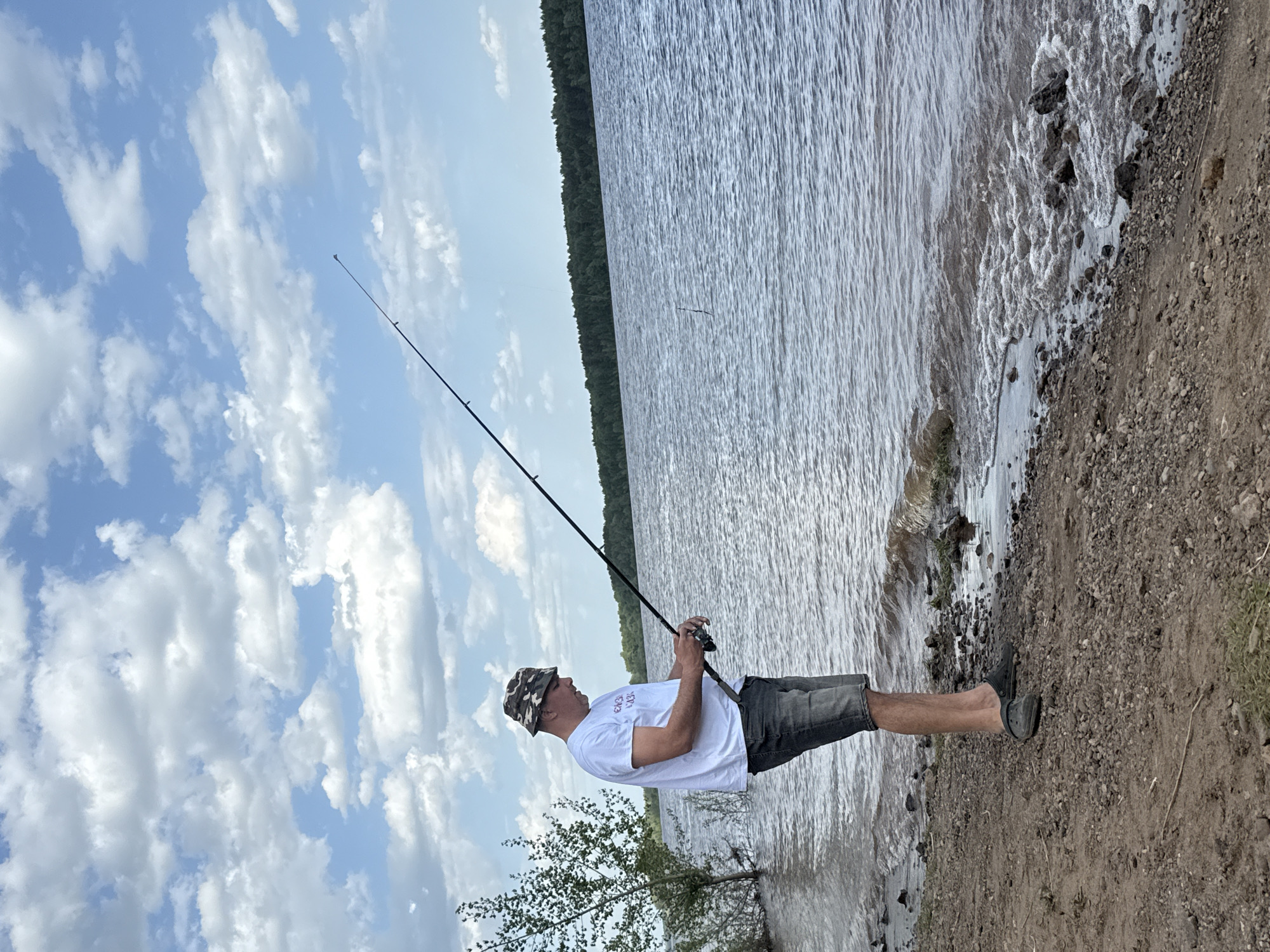 A man is fishing at Doksha Lake in Doksha, Russia. The man is wearing a white t-shirt, grey shorts, a camouflage bucket hat, and black shoes. He is holding a fishing rod. The lake is in the background, with a shoreline covered in pebbles. The sky is blue with white clouds.