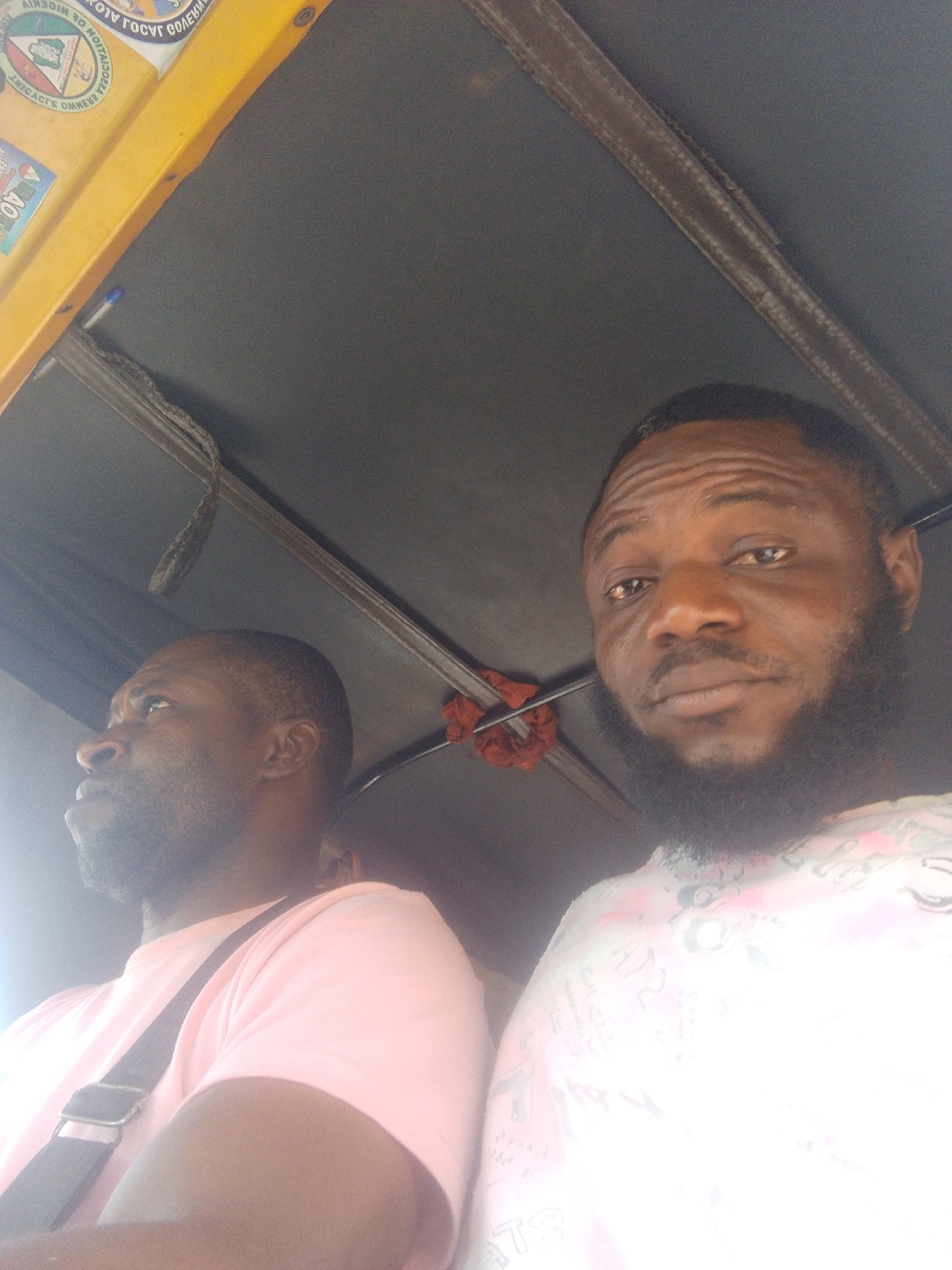 Two dark-skinned men are inside a vehicle, likely a taxi or auto rickshaw, in the city of Unknown, Unknown. The man on the left wears a pink t-shirt with a strap over his shoulder, looking towards the left. The man on the right has a full beard and is looking directly at the camera, wearing a white t-shirt with writing on it. The vehicle has a grey interior roof, with visible supports and attachments. Stickers are seen in the upper left corner.