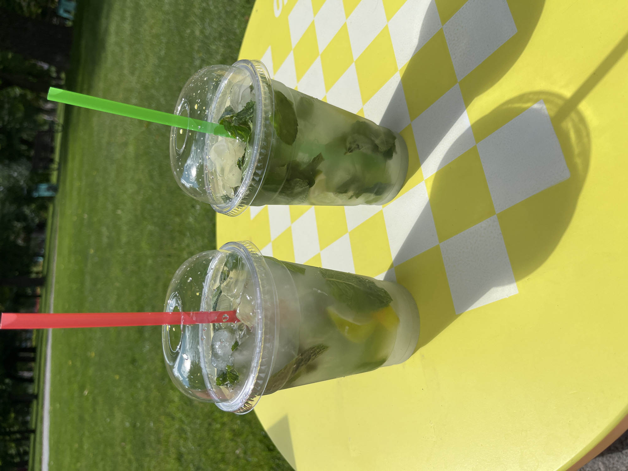 Two clear plastic cups filled with a light-colored beverage, ice, mint leaves, and lime wedges sit on a bright yellow table with a white checkered pattern. Each cup has a clear plastic lid and a colored straw protruding from it; one straw is red, and the other is green. The cups appear to be refreshing drinks, possibly mojitos. The table is outdoors, with a grassy area and trees visible in the blurred background under a sunny sky. The image is framed from a slightly elevated perspective looking down at the table. The setting is likely a park or outdoor seating area in Kharkiv, Ukraine, suggested by the context of recent requests and similar imagery. There are no people visible in the scene. The sunlight casts distinct shadows from the cups and straws onto the checkered table surface.