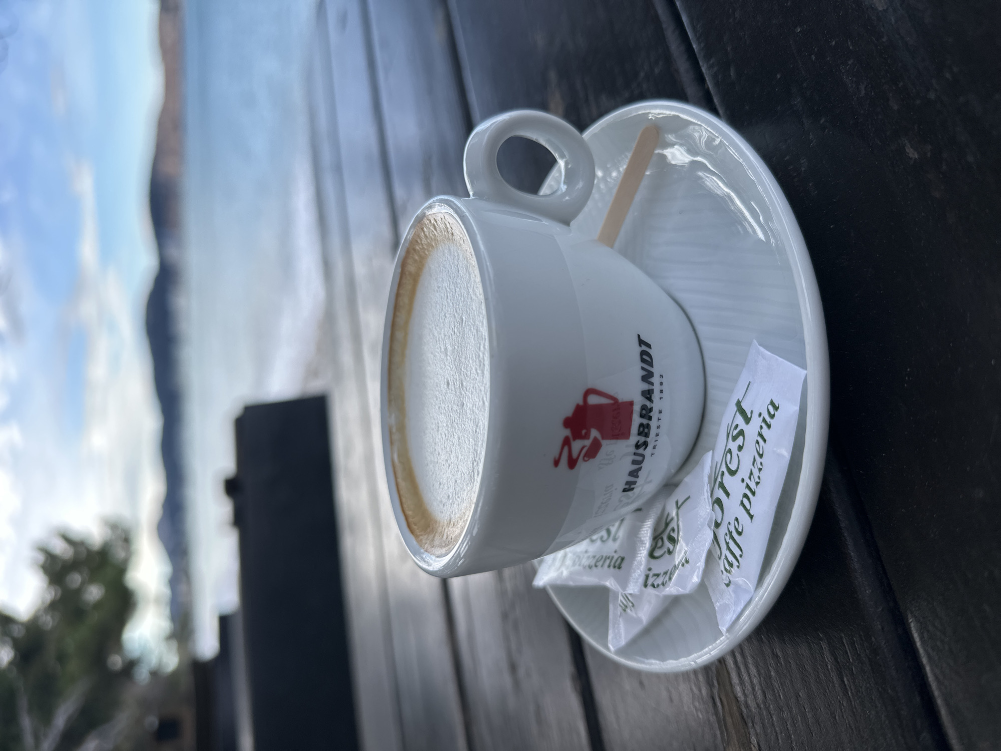 A cappuccino in a white cup rests on a white saucer on a dark wooden table in Šušanj, Montenegro. The cup has a red logo that includes the words "HAUSBRANDT" and "TRIESTE 1892". Several sugar packets labeled "Forest, Caffe Pizzeria" are on the saucer. A wooden stirrer is also on the saucer. In the background is a blurred view of the coastline and mountains, typical of the Šušanj area. The image is taken at a close range, emphasizing the coffee and table setting.