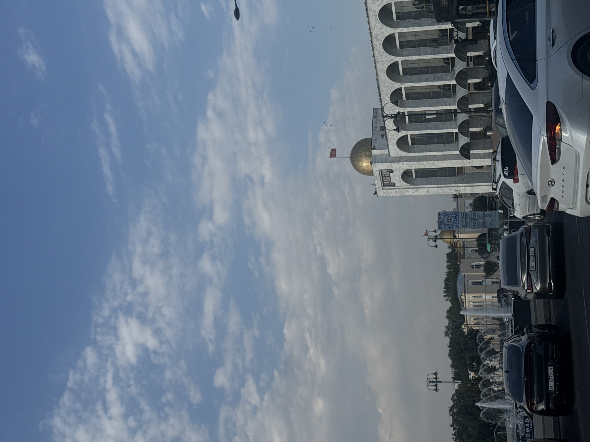 The image depicts a street scene in Bishkek, Kyrgyzstan, featuring a prominent building with a golden dome and arched windows, likely a government or cultural institution. In the foreground, several cars are parked and in motion, indicating traffic. Fountains are visible in the background, suggesting a public space or park. The sky is partly cloudy, with a clear blue expanse overhead. The overall impression is that of a bustling urban environment on a clear day.