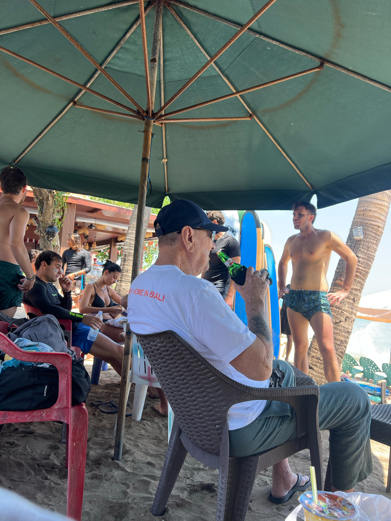 The image depicts a group of people gathered on a beach in Kuta, Indonesia, under a large green umbrella providing shade. In the foreground, an elderly man wearing a black baseball cap and a white t-shirt with "MY HOME IN BALI" printed in red is seated, holding a green bottle in his right hand. To his left, a younger man in a black wetsuit and blue shorts is seated, looking down. A woman in a black bikini is seated next to him, holding a bottle of water and a food item. Behind them, several surfboards are propped upright. To the right of the scene, a young man wearing patterned swim shorts stands with his hands on his hips, facing away from the camera. The background shows a beach with sand, palm trees, and ocean waves breaking on the shore. The overall environment suggests a casual beach setting with people enjoying leisure time.