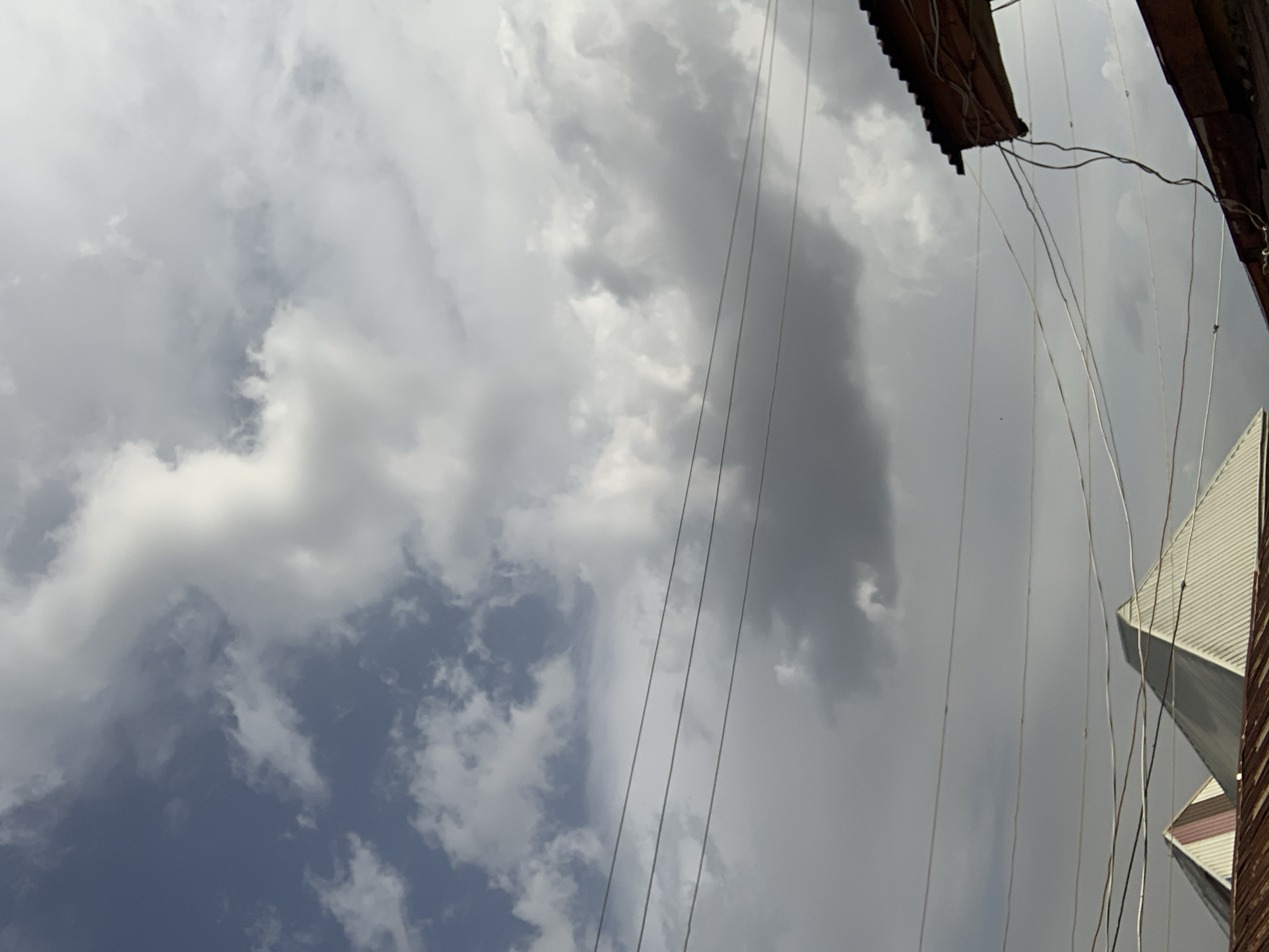 A view of a cloudy sky with power lines running across the frame. Building edges and roofs appear in the top right and bottom right of the frame. The image could be from Rafin Par, Nigeria.