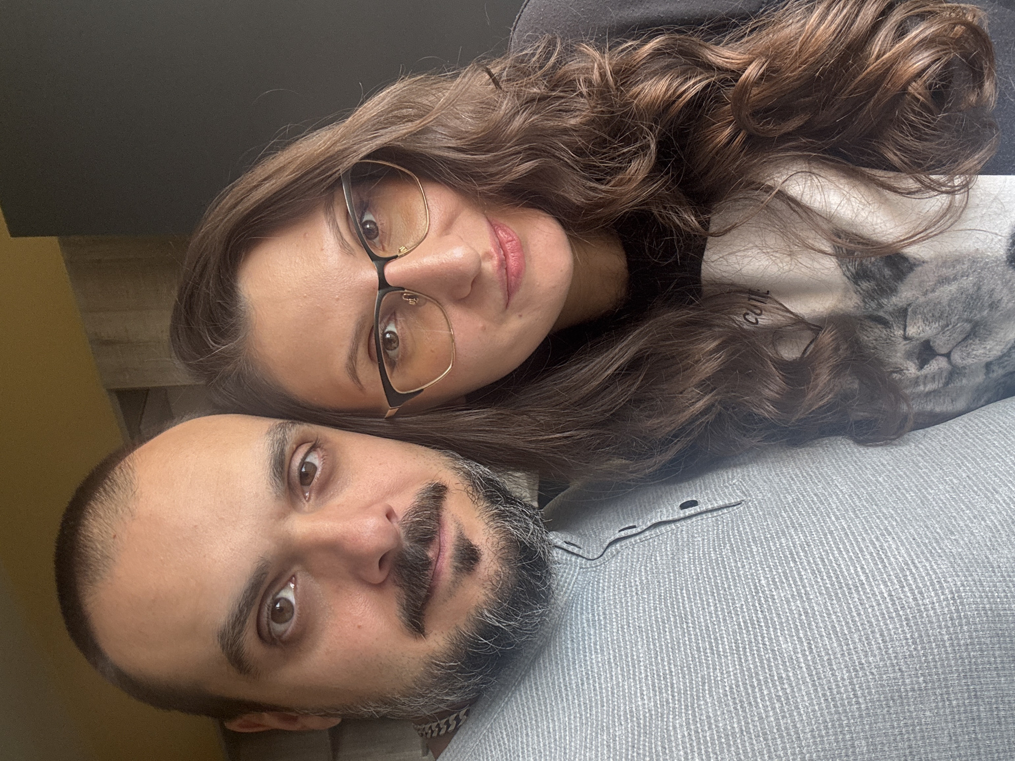 A close-up shot features a woman and a man. The woman, positioned at the top, has long, wavy brown hair and wears glasses with gold frames. She is wearing a black top with a white cat print. Her expression is serene, and she appears to be looking directly at the camera. The man, below the woman, has short, dark hair and a graying beard. He is wearing a gray, buttoned shirt. The image is shot indoors, with what appears to be a gray background partially visible. There is no information to infer a location in Sofia, Bulgaria.