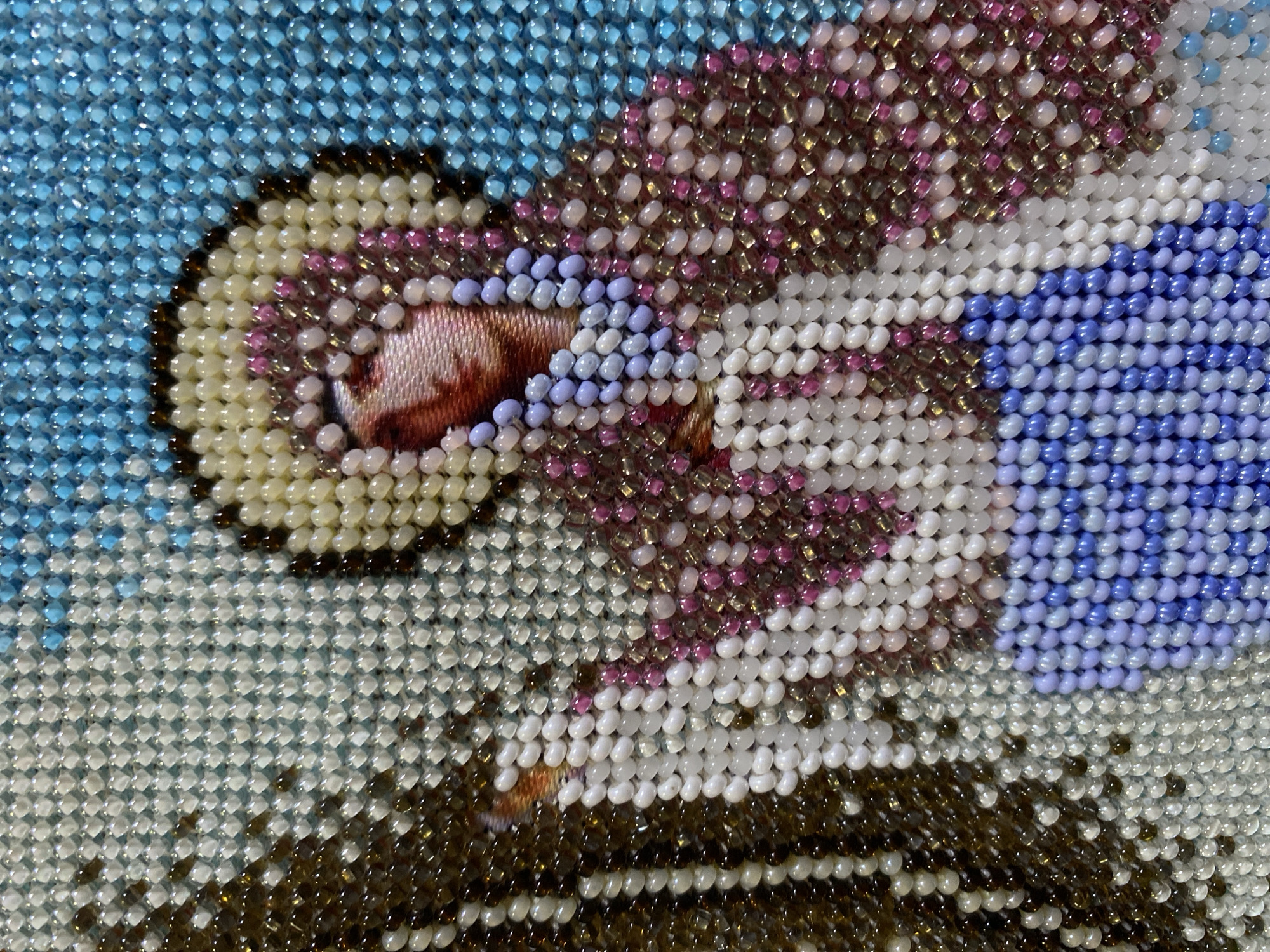 This is a close-up, macro photograph of a beaded tapestry or artwork. The subject appears to be a religious figure, possibly a saint or an angel, rendered in a stylized manner using a mosaic of small, colored beads. The figure has a halo made of cream-colored beads and a face depicted with flesh-toned and pink beads. The figure is wearing a robe that transitions from white and pink to a vibrant blue at the bottom, with accents of purple and gold beads. The background is a mosaic of sky-blue beads. The artwork is highly detailed, with each bead meticulously placed to form the image. There is no discernible information about its location in Zgrade, Montenegro, as the image solely focuses on the texture and detail of the beadwork.