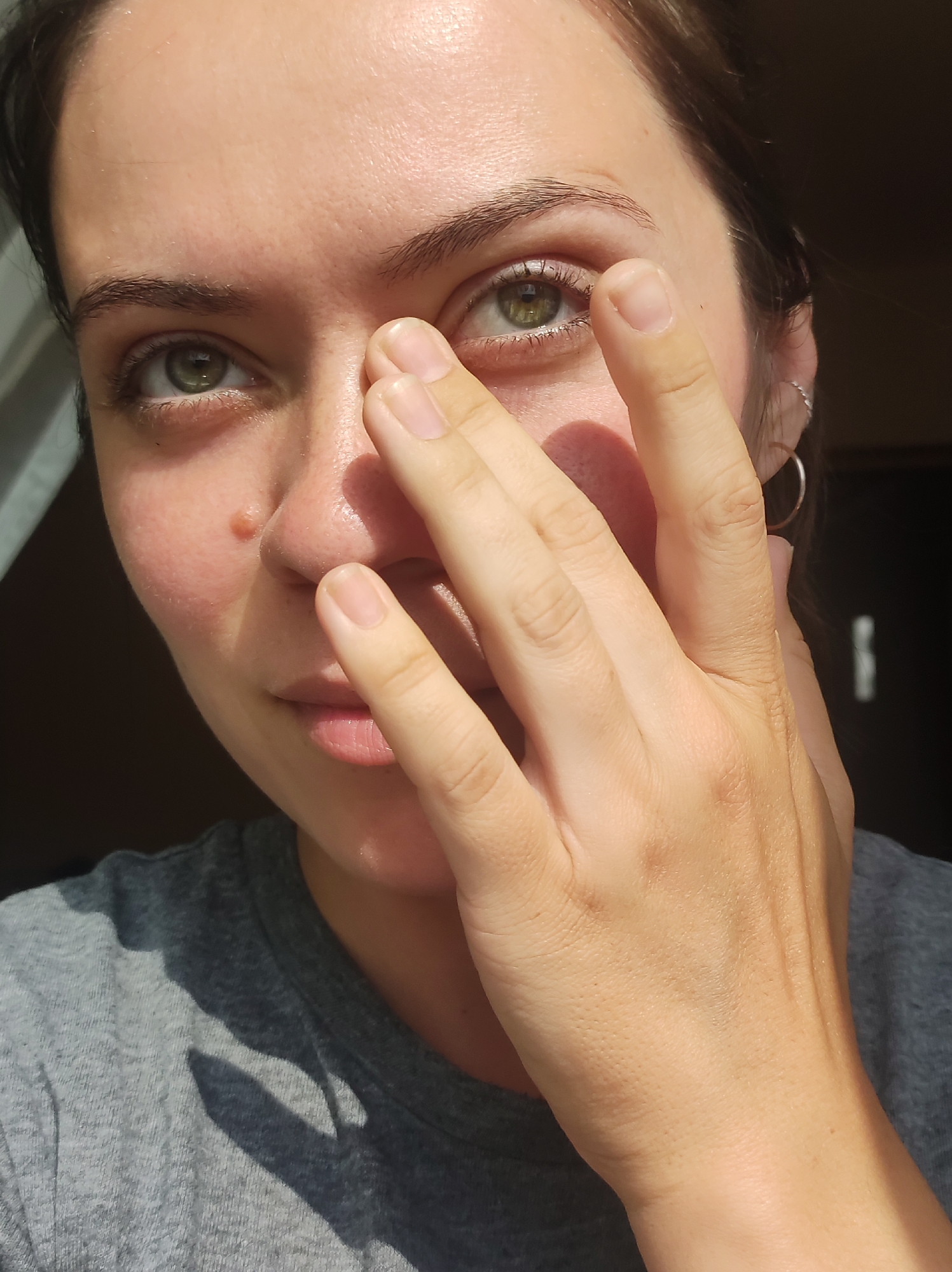 A close-up, outdoor, eye-level shot shows a woman's face, partially obscured by her hand, in Chaikovsky, Russia. The woman has green eyes and her right eye is visible as she touches her face with her right hand. Sunlight illuminates her face, casting shadows across her skin and her gray t-shirt. She has a small mole on her nose and is wearing a silver hoop earring. The background is dark, suggesting an indoor setting or a shaded area.