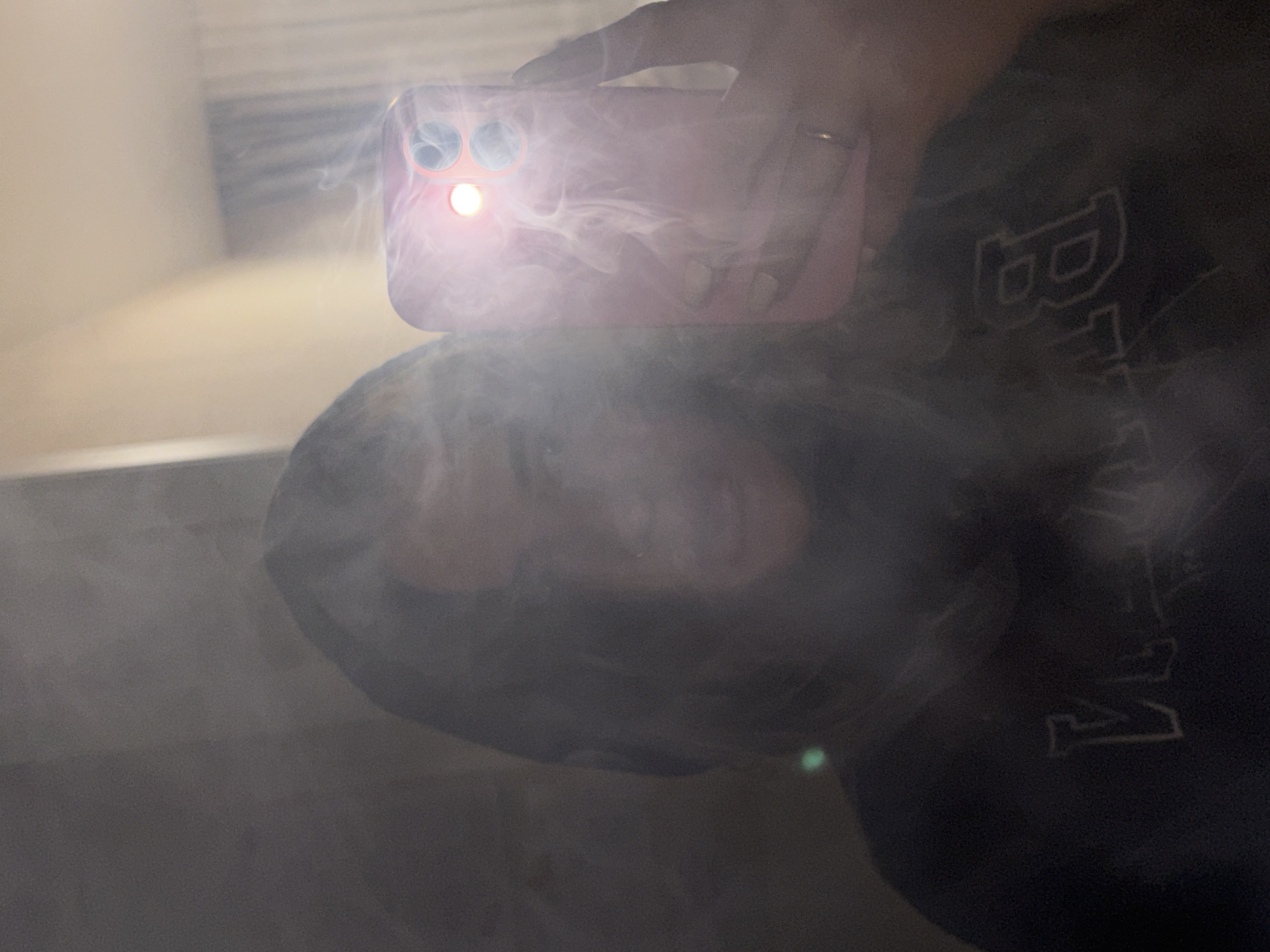 The image displays a self-portrait captured through a reflection, likely from a mirror or reflective surface. A person is holding a pink/red smartphone with their hand, which wears a ring. The smartphone is highly visible in the foreground, featuring three camera lenses and a bright, active flash that emits intense light.
This powerful flash illuminates a dense, swirling cloud of what appears to be smoke, steam, or vapor, which creates dynamic light patterns and partially obscures the reflected scene. Behind the phone and the haze, the person's reflection is visible, showing dark hair and the upper portion of their face, largely veiled by the bright light and vapor.
The individual is wearing dark clothing, possibly a hoodie or sweater. On this garment, a large, stylized letter "B" is clearly visible, outlined in a lighter color, followed by other indistinct, outlined characters. The background of the reflection suggests an indoor setting with a light-colored wall. The use of the flash implies low light conditions. A small green lens flare is also noticeable in the lower part of the reflection. The overall effect is a luminous and slightly mysterious composition, with the light from the phone playing a central role in interacting with the surrounding vapor. The location context provided is Krasnodar, Russia.