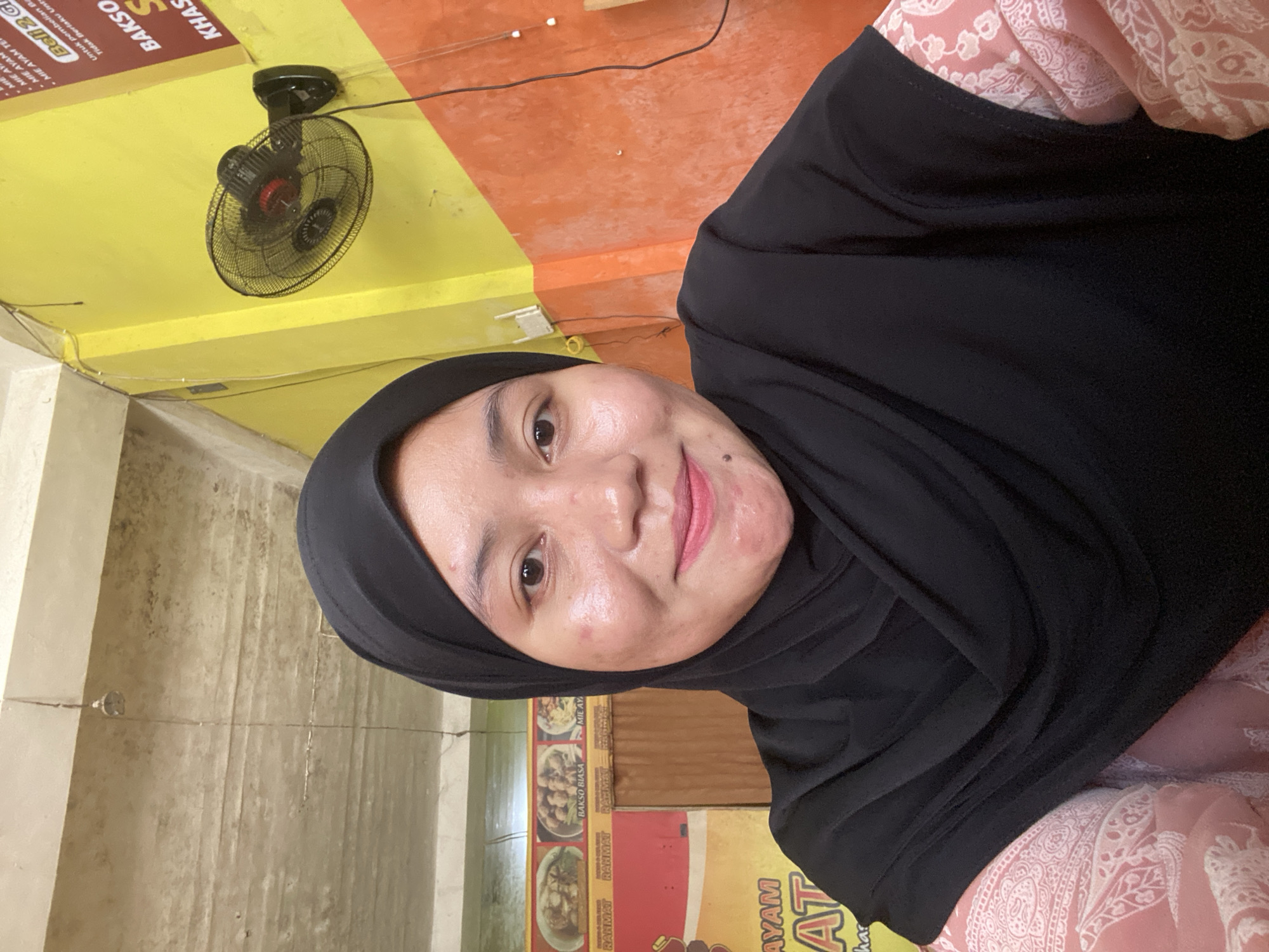 A young woman wearing a black hijab and a pink patterned shirt smiles at the camera in what appears to be a restaurant. The background features yellow and orange walls, a ceiling fan, and signs in Indonesian. One sign shows images of food, with text including "RAMAT" and "BAKSO BIASA". Another sign reads "AYAM" and "AT". The ceiling is concrete with visible cracks and wires. The overall impression is of an indoor dining establishment in Sungai Sipai, Indonesia.