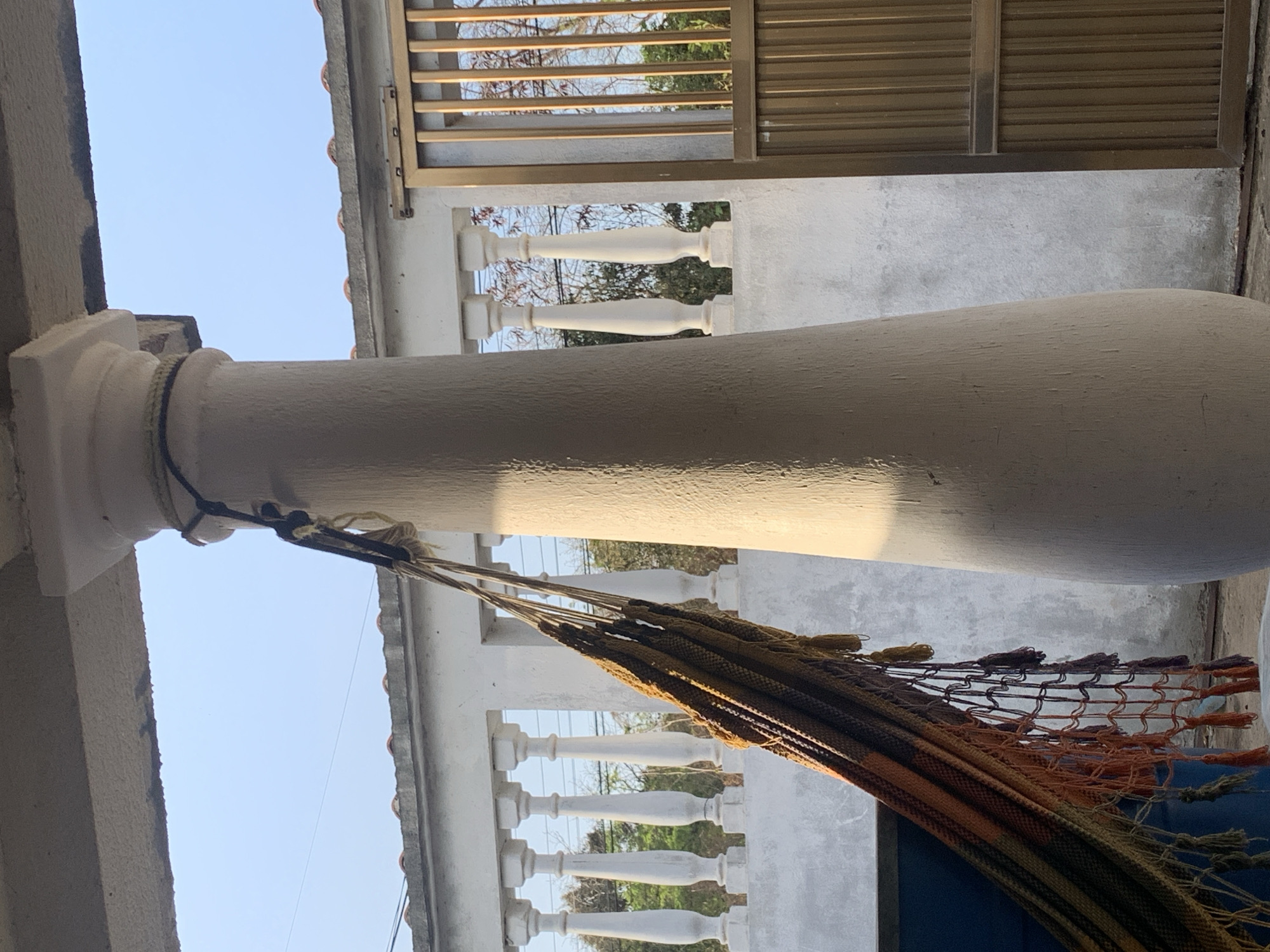 A low-angle, slightly rotated view looking upwards at a white column supporting a structure. A hammock hangs from the column, its netting and fringe details visible. In the background, a balustrade with white posts is present, allowing a glimpse of trees and sky. The sky is pale blue. A slatted metal structure is partially visible at the top. The location is suggestive of an outdoor space, possibly a patio or balcony, but the specific location in Santa Ana, Venezuela cannot be determined from this image alone.